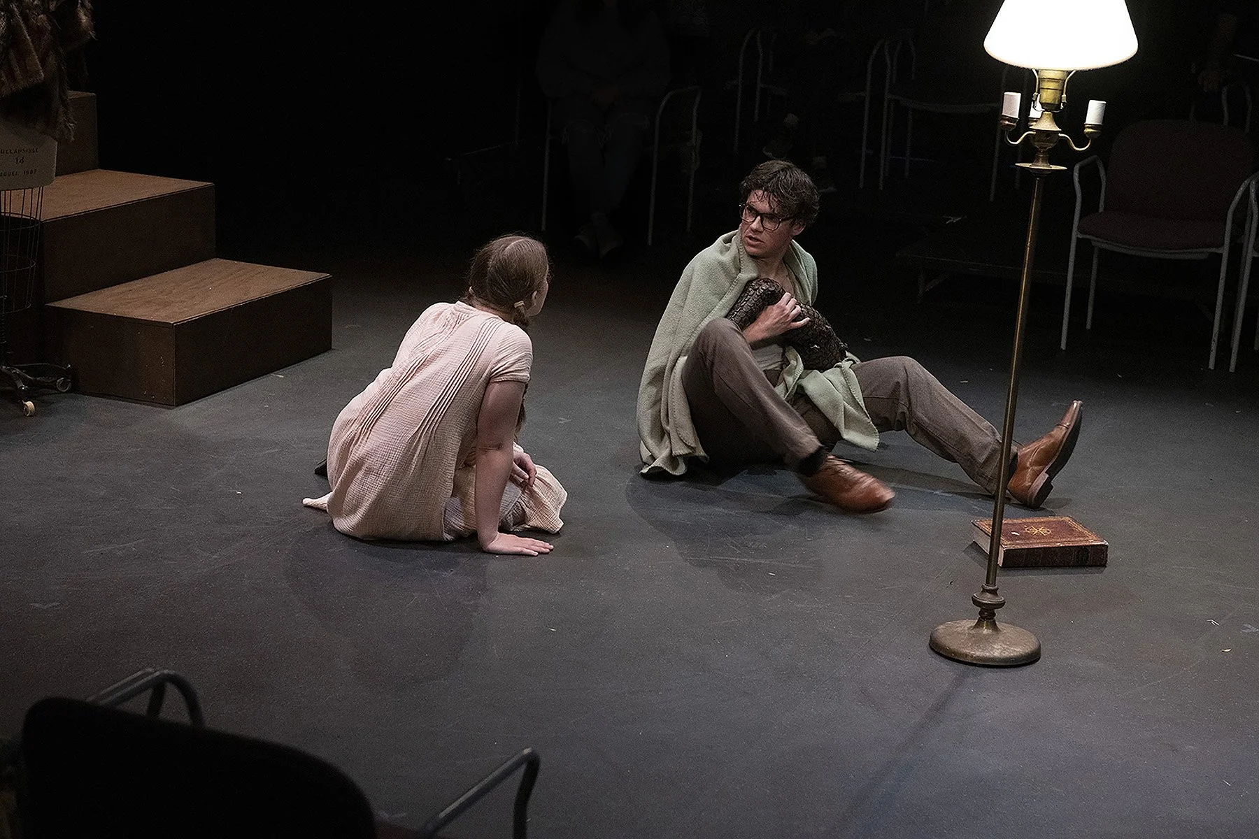 Two actors on stage during a theatrical performance. One actor, a woman, is kneeling and wearing a light-colored dress, while the other, a man, is sitting on the floor with a cardigan draped over his shoulders, wearing glasses, brown pants, and boots