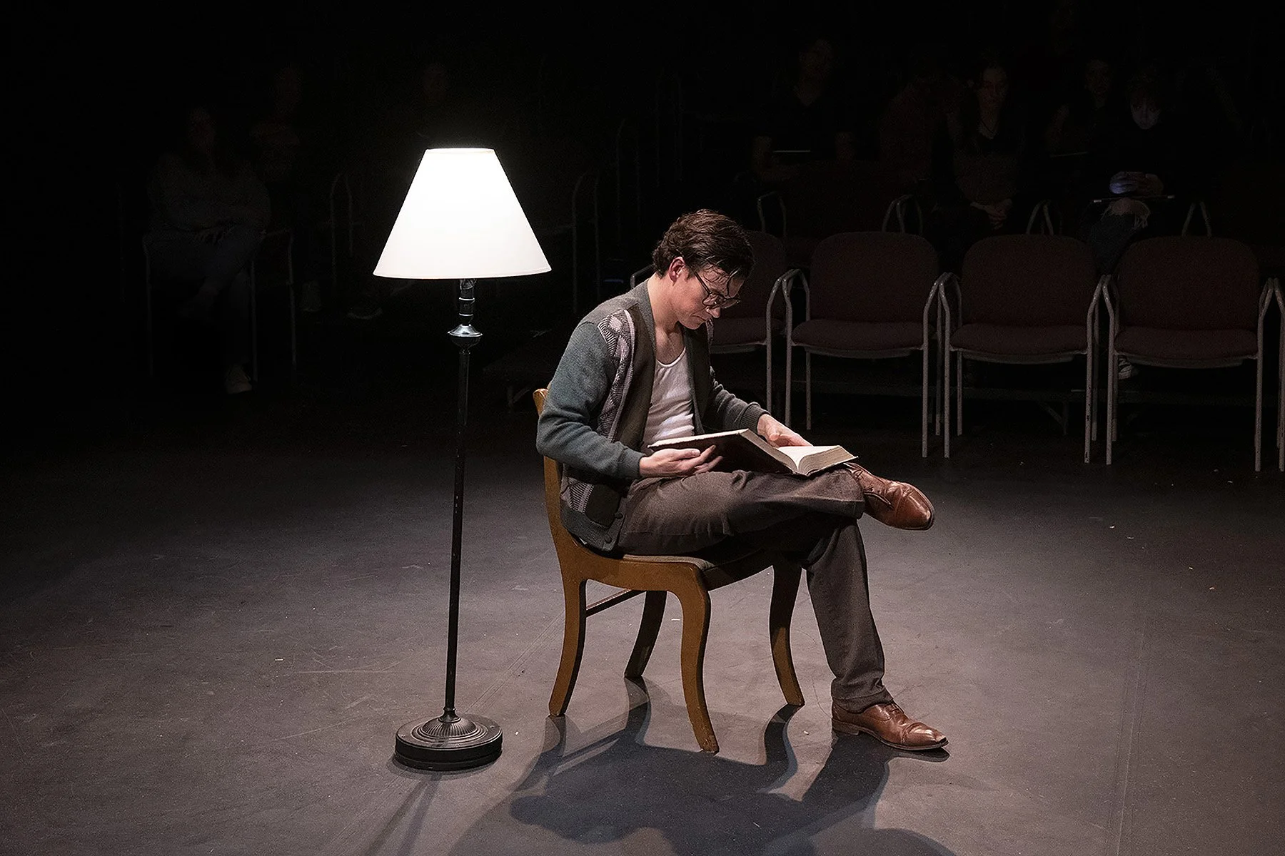 A person sitting on a wooden chair in a dimly lit space, reading a book. A standing lamp provides light. Empty chairs are visible in the background.