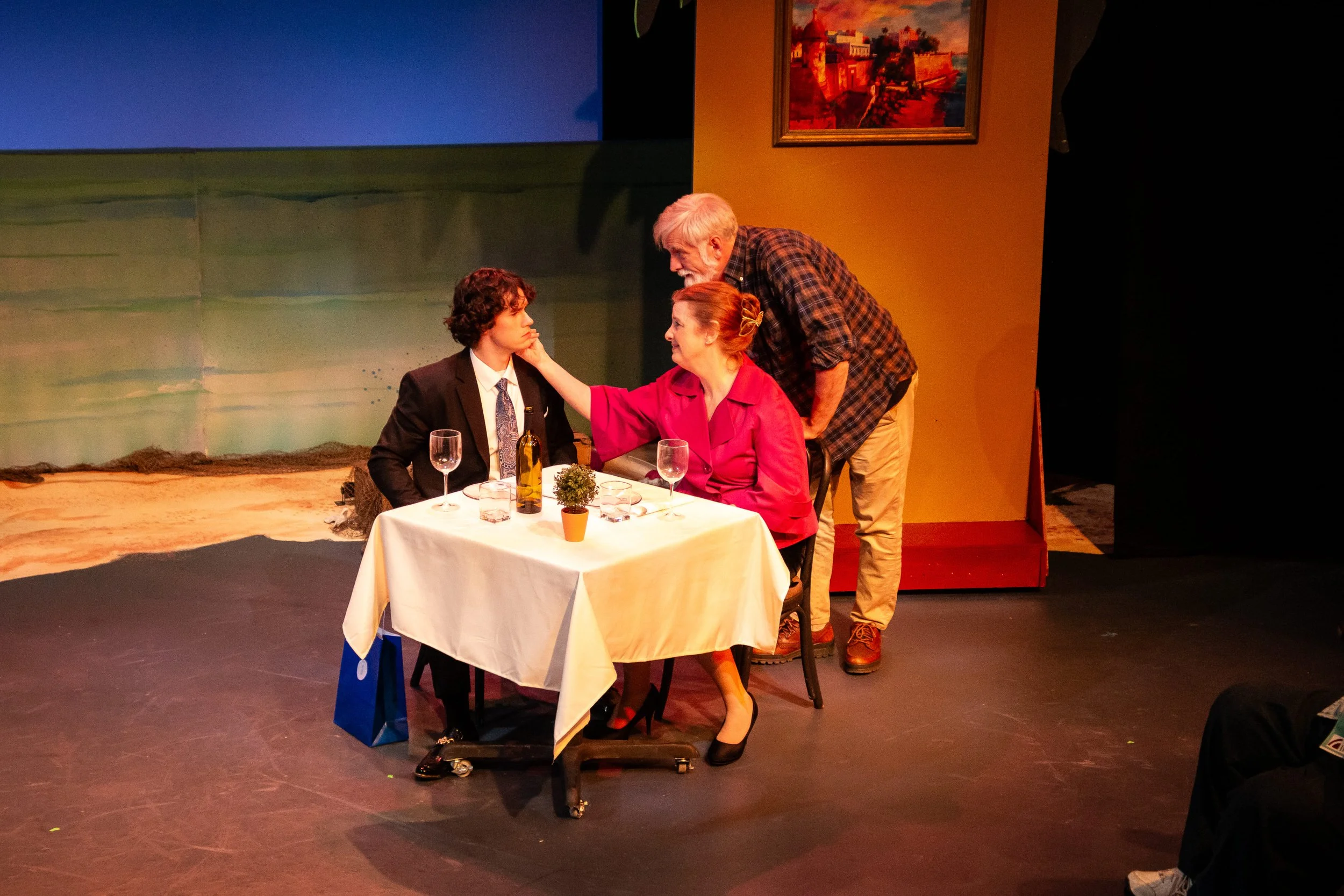 Stage scene from a play with three actors. A young man in a suit and a woman in a pink dress are sitting at a table, with an older man leaning over talking to the woman. The table has a wine bottle, two wine glasses, and a small potted plant. The bac