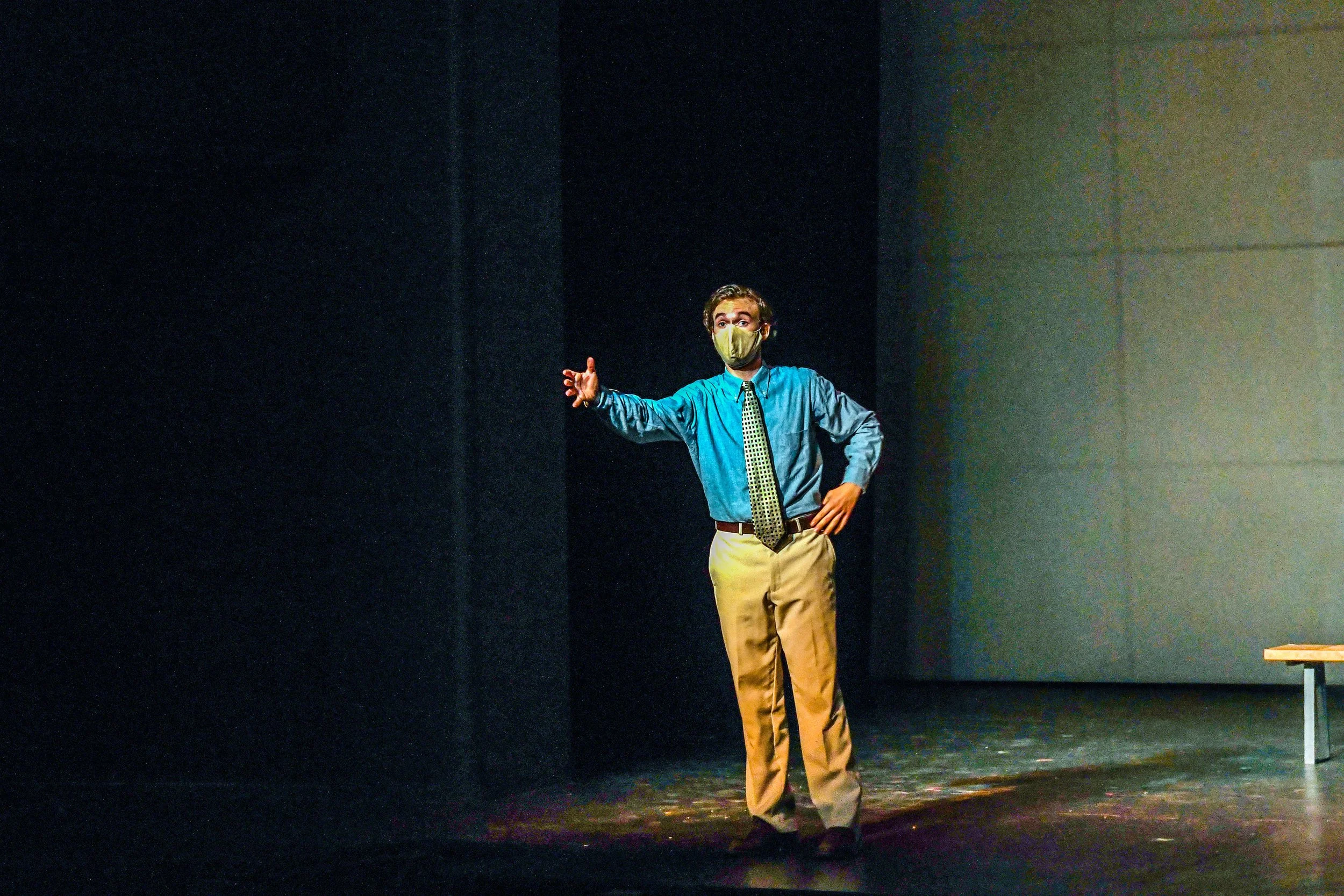 A man standing on a stage wearing a mask, light blue shirt, and tan pants, gesturing with one hand, dark background, with a small table to the right.