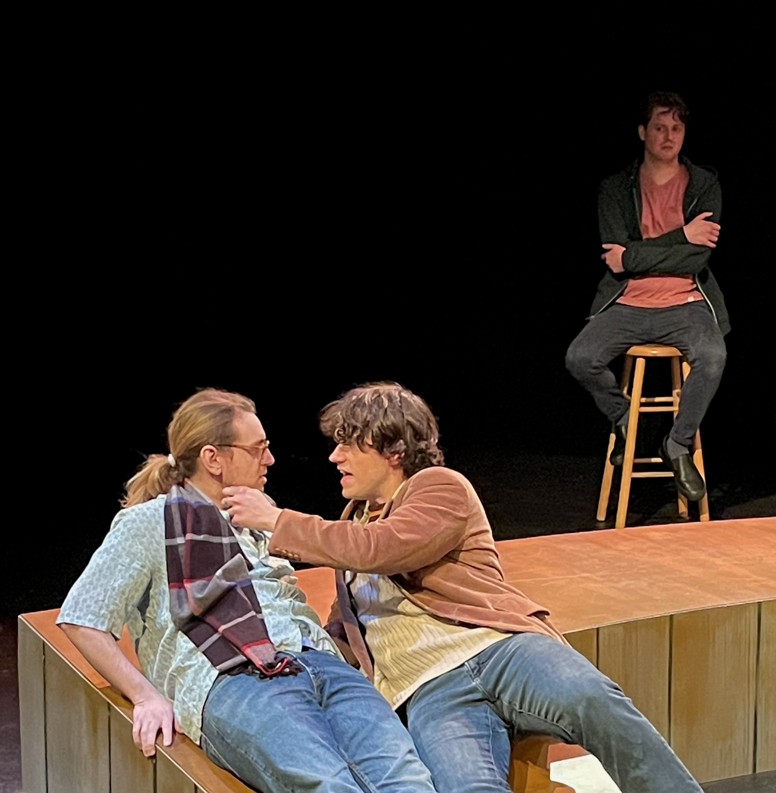 Two men sitting closely at a wooden table, engaging in conversation with one touching the other's shoulder, in a warm, intimate setting.