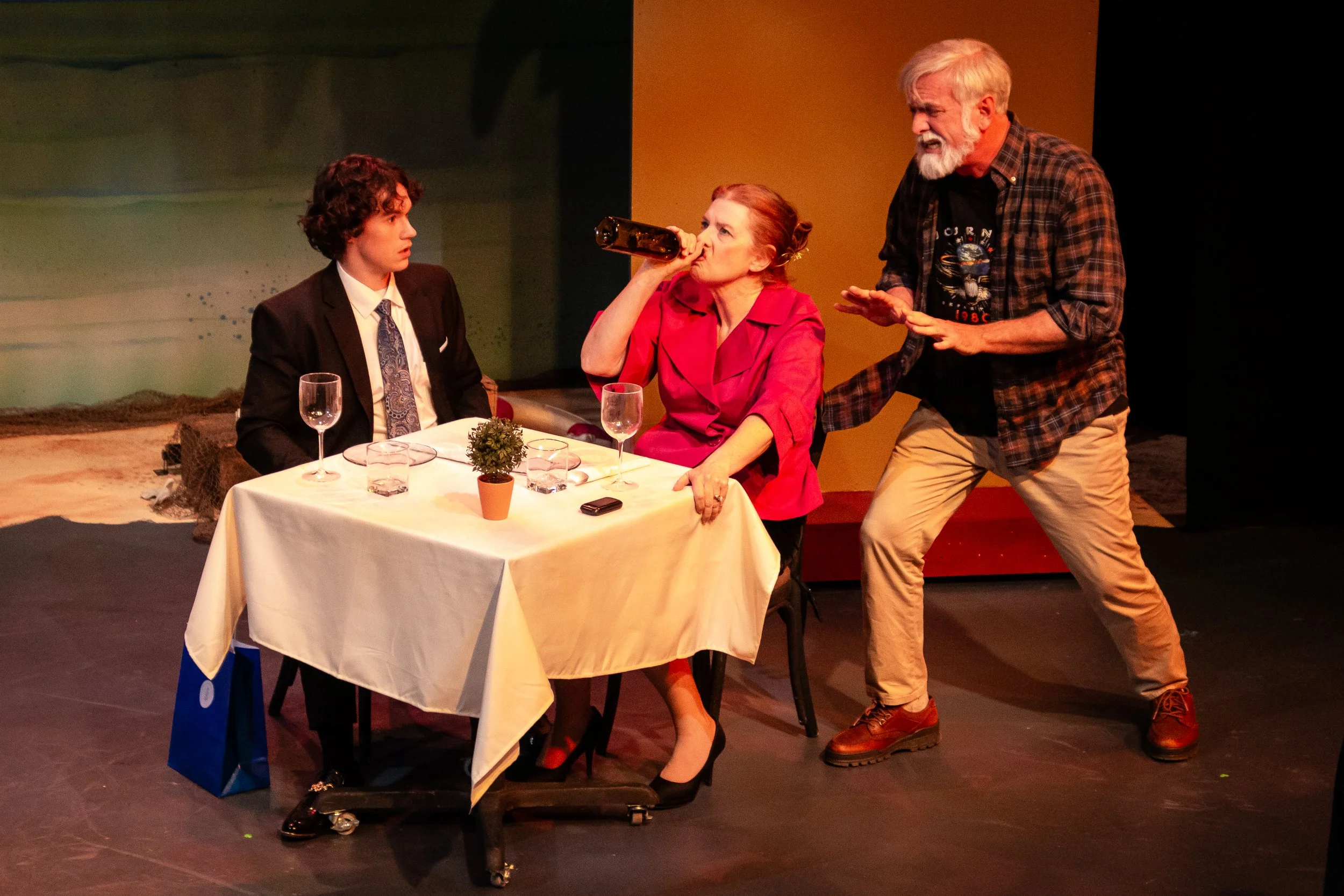 A theatrical scene with three actors. A young man in a suit and tie sits at a table, looking surprised. An older woman with red hair and red dress is drinking from a bottle. A bearded man in casual clothes is standing and gesturing passionately.