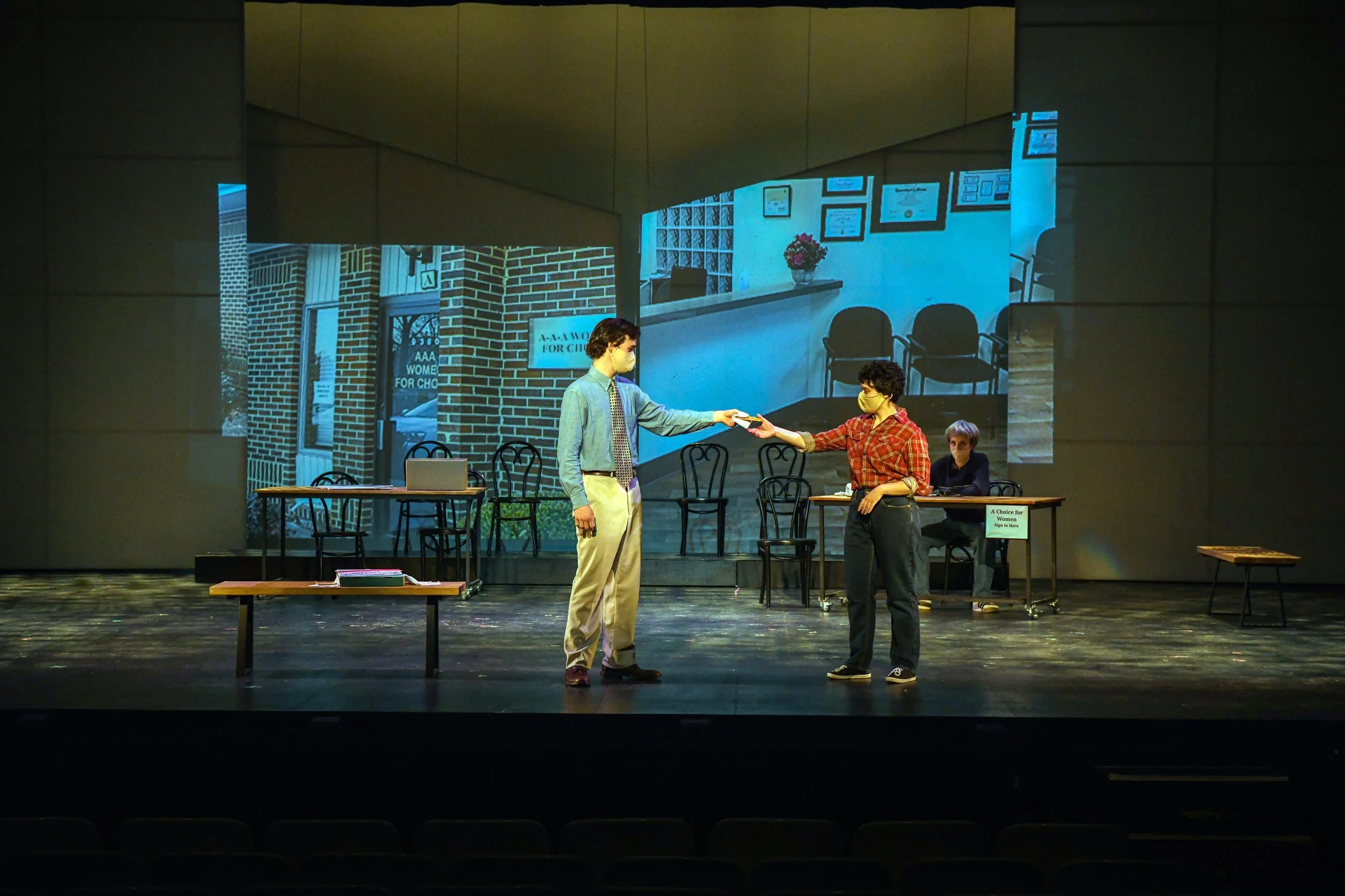 The image depicts a stage performance with three actors. One actor, dressed in a light blue shirt and beige pants, is handing something to a second actor wearing a red plaid shirt and dark pants. The third actor, seated at a desk in the background, i