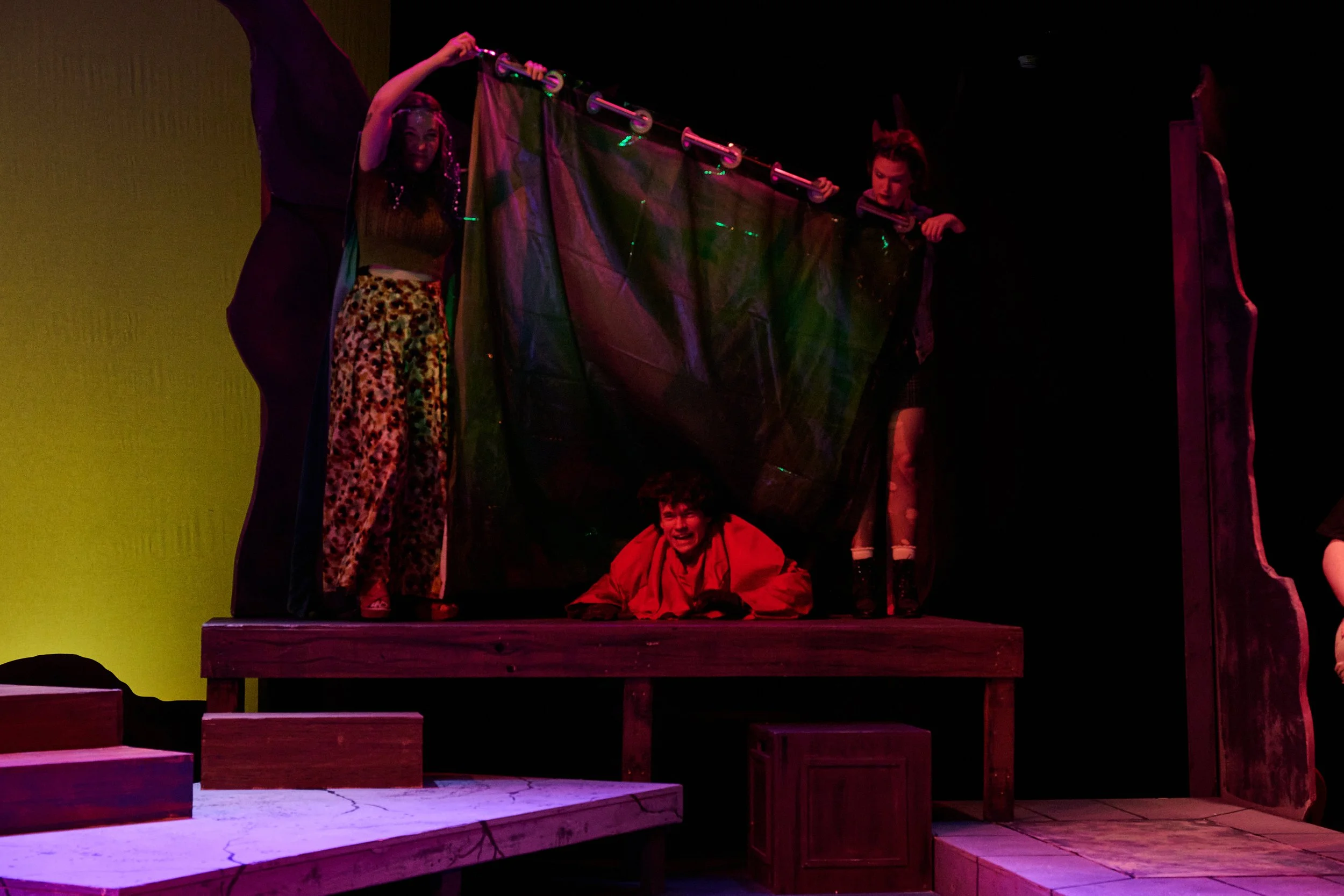 Group of five people on a stage, some holding a fabric backdrop, one person sitting on the floor in red clothing, the background is dark with colorful stage lighting.