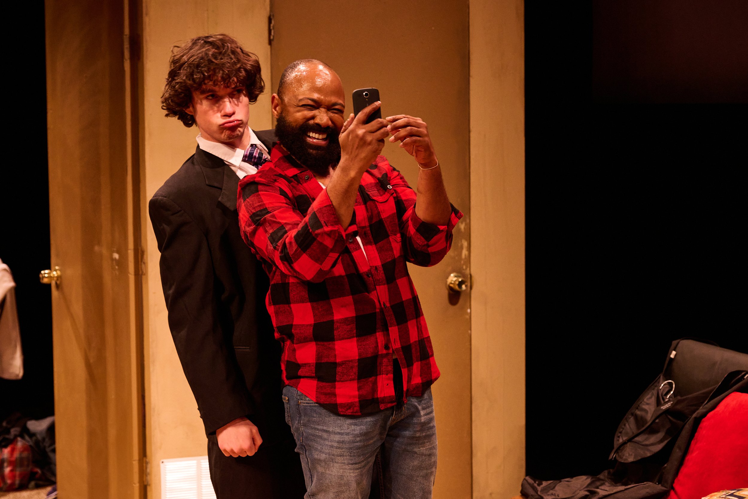 Two men taking a selfie together. One man has curly brown hair, a light complexion, and is wearing a dark jacket over a white shirt with a tie. The other man has a darker complexion, a beard, and is wearing a red and black checkered shirt, smiling wi