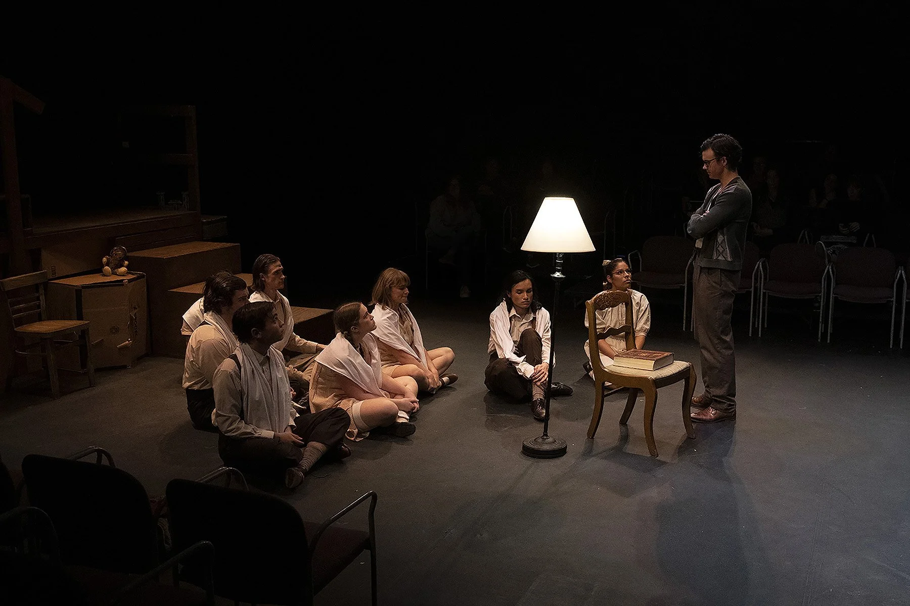 A theatrical scene with seven people sitting cross-legged on a dark stage, facing a man who is standing. The man is wearing glasses and a gray sweater, with his arms crossed. The stage has a small table with two books on it, a lamp, and some props li