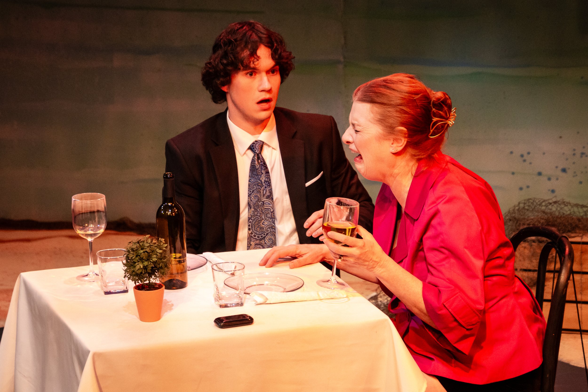 A young man in a suit and an older woman in a red jacket are sitting at a dining tablet. The young man is shocked and concerned because the woman is crying. She is also holding a glass of white wine.