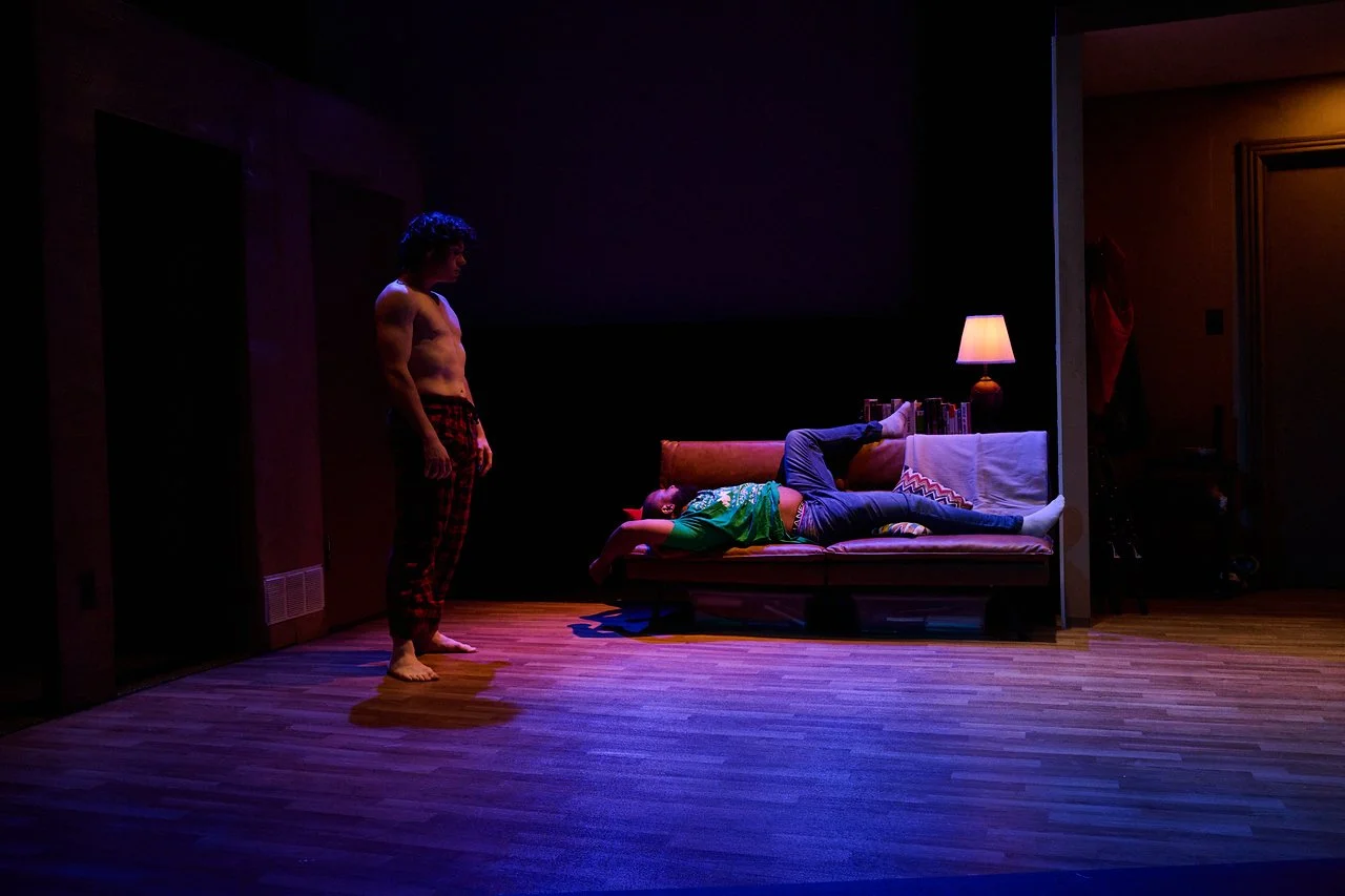 A shirtless young man stands on a wooden floor facing a woman lying on a sofa. The woman is wearing jeans and a green top, and is lying on her side with one leg raised. The scene is dimly lit with a lamp on a table next to the sofa, creating a warm g