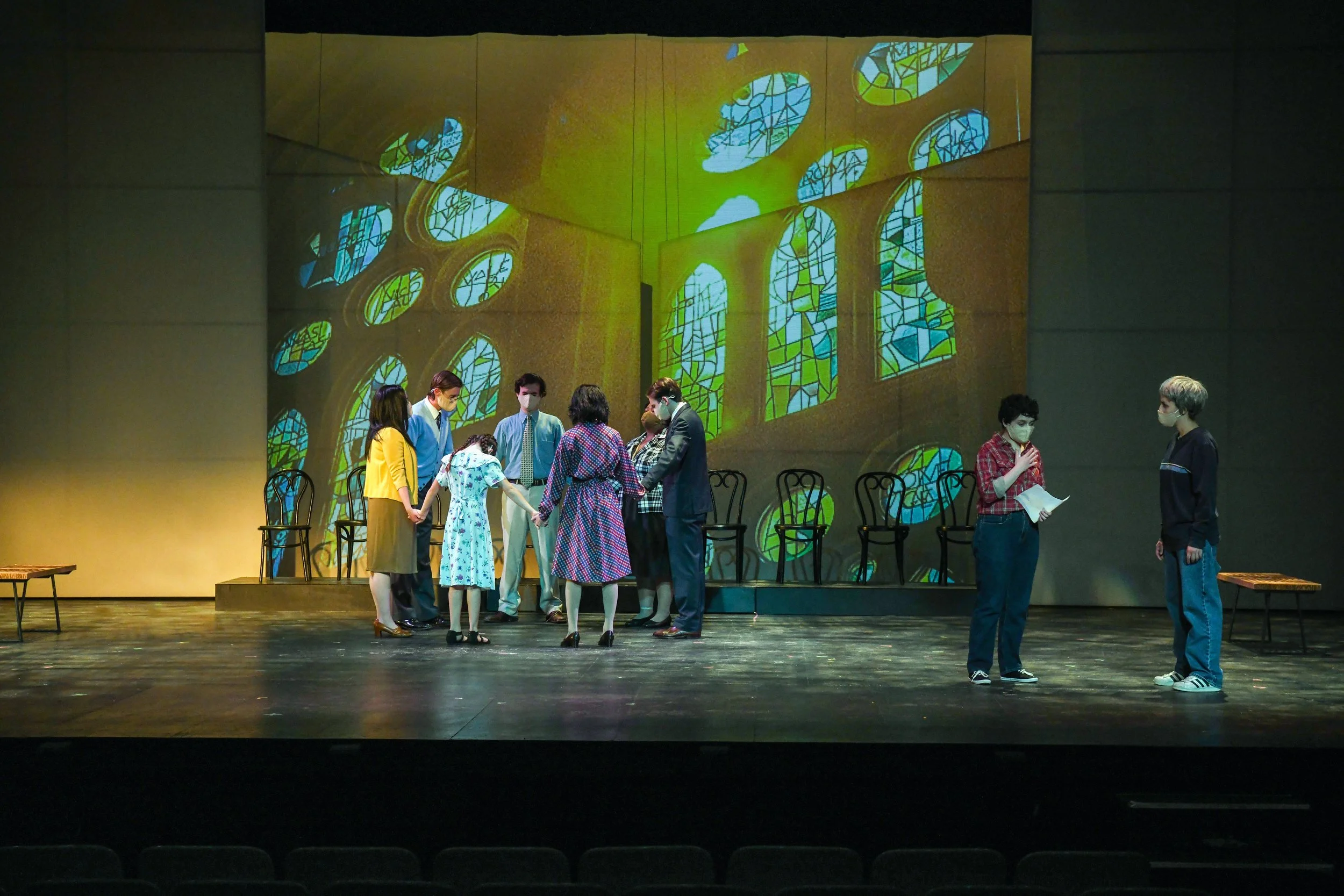 The photo shows a theater stage with a stained glass window projected onto the back wall. Several actors or performers are standing in a group in the center of the stage, some holding hands and engaged in conversation or a scene. There are empty chai