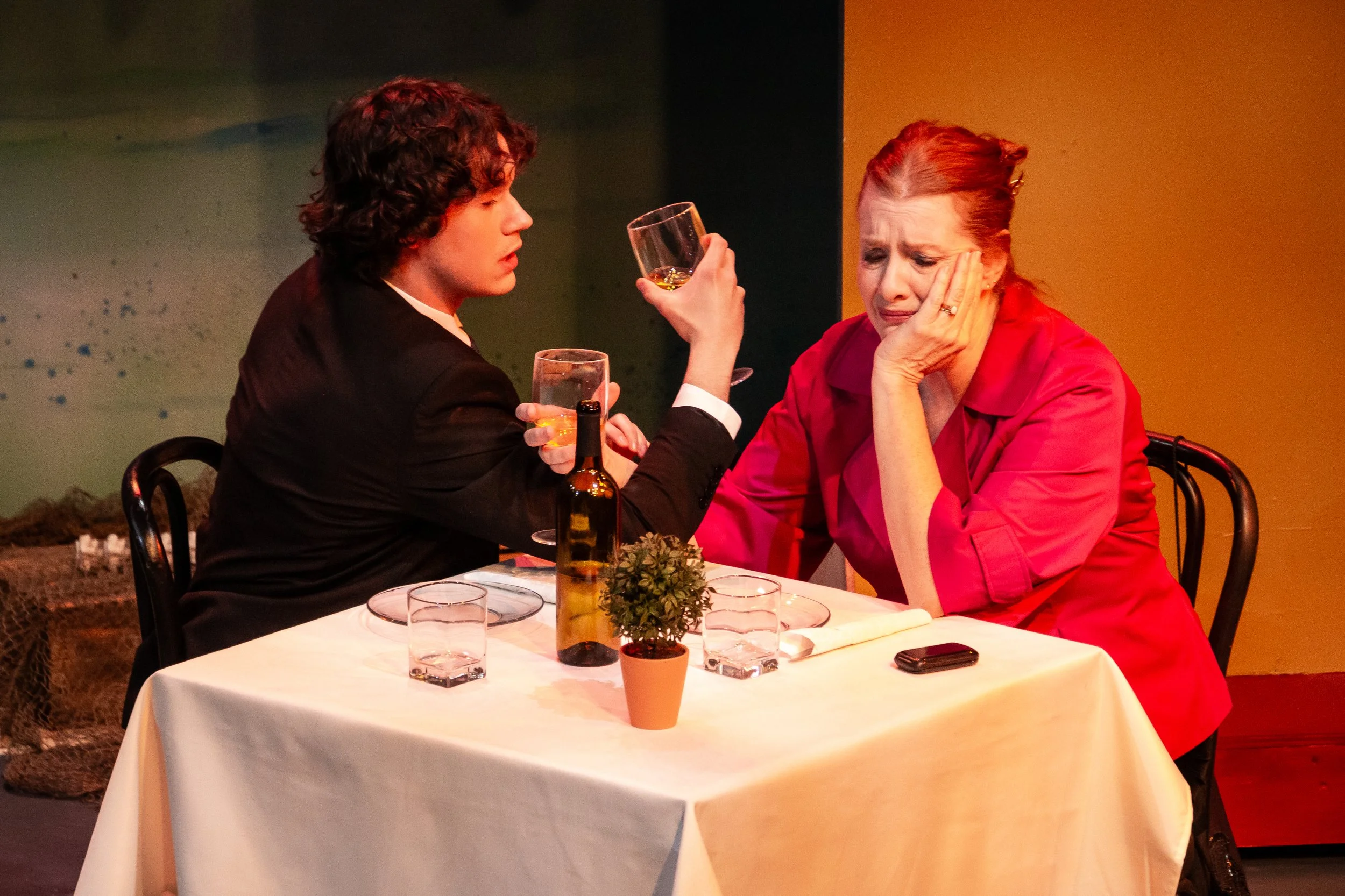 A young man with curly hair and a woman with red hair sit at a dinner table, with the man appearing to console or listen to the woman, who looks distressed. The table has glasses, a small potted plant, and a bottle of beer.
