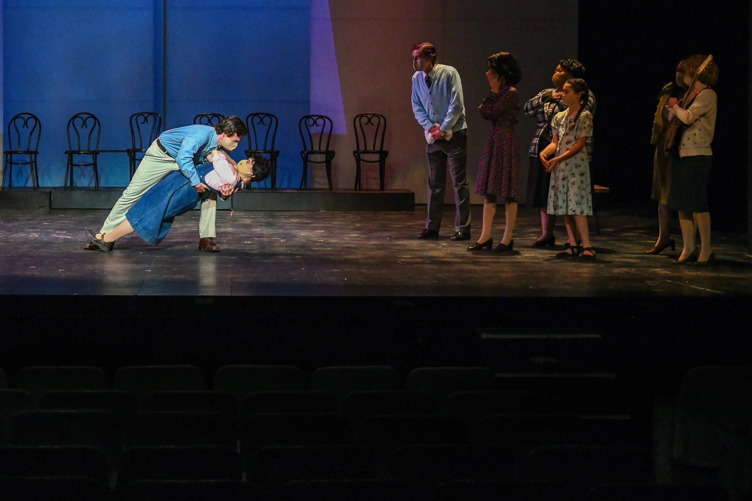 A group of people on stage, with one person bent over and holding another person, while others watch. The stage has empty chairs in the background.