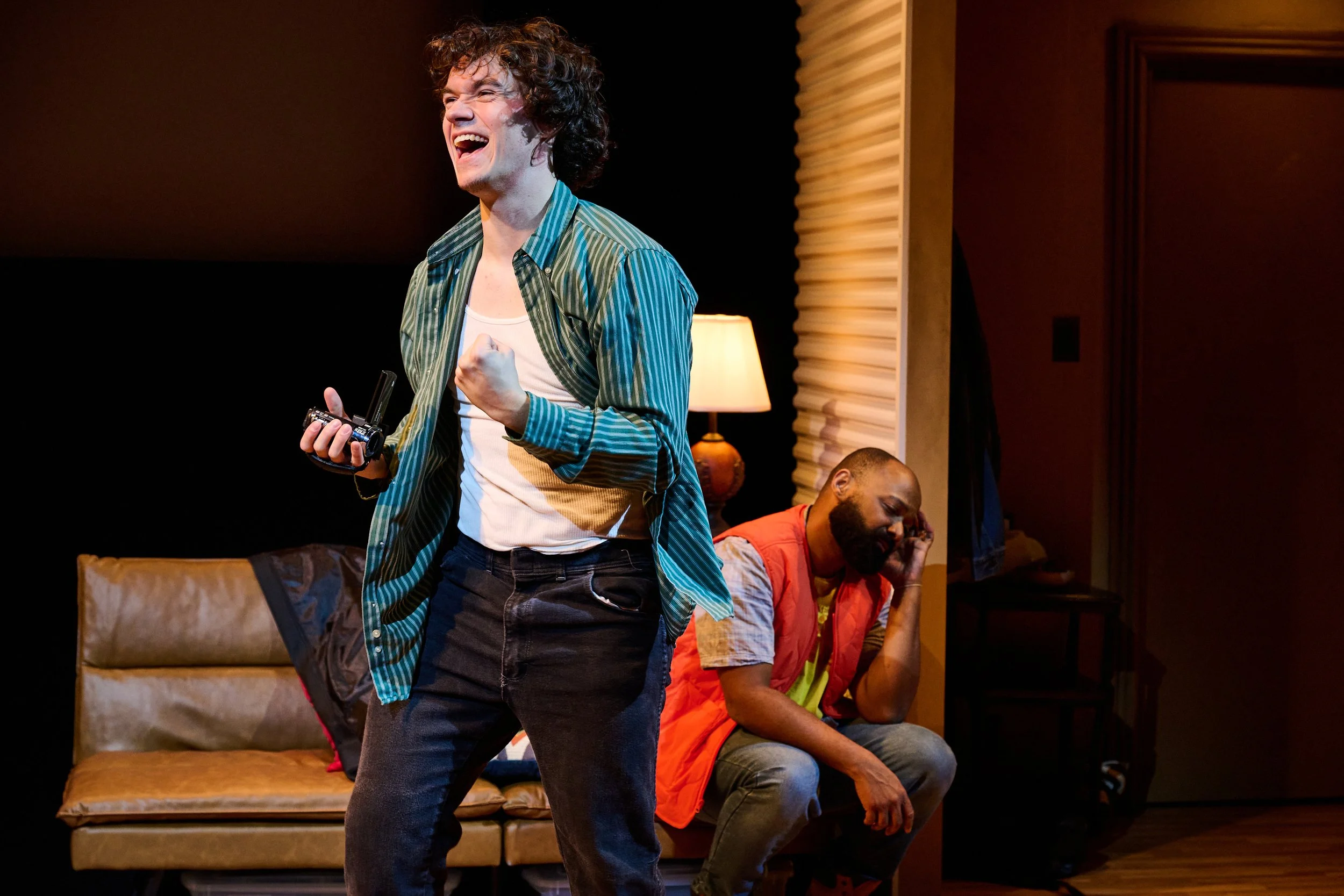 A man with curly hair in a green striped shirt is laughing and celebrating, holding a walkie-talkie, while two other men sit on the couch behind him, one of them with a beard and wearing a red vest, looking down.