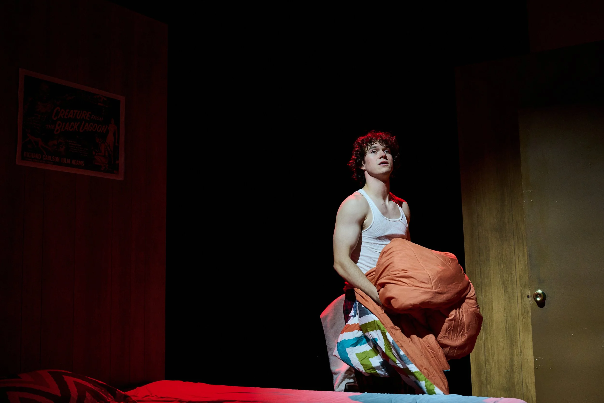 A person with curly hair wearing a tank top sitting on a bed, with a dark room and a poster on the wall that reads 'Creature from the Black Lagoon'.