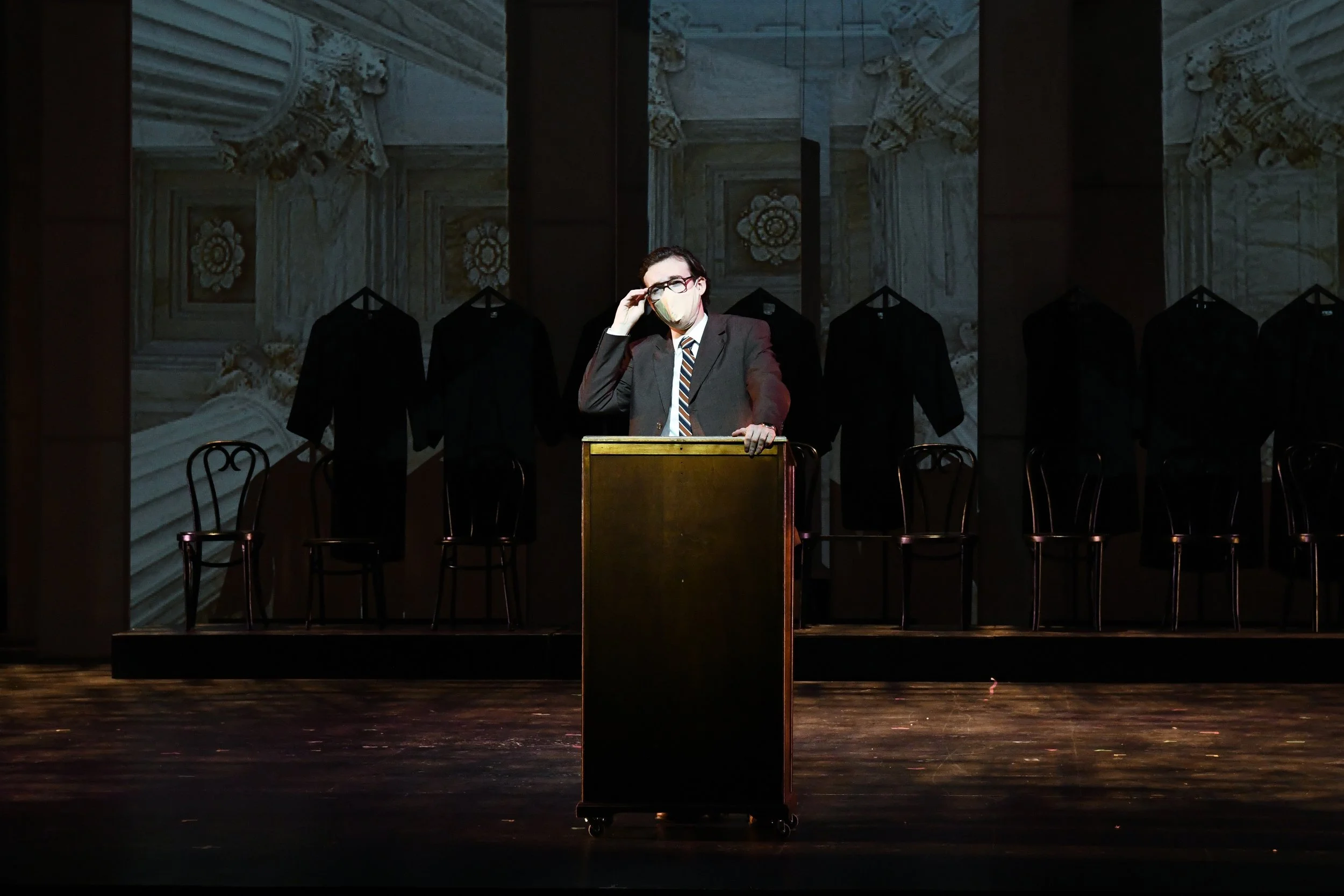 A man in a suit and glasses wearing a face mask stands behind a podium on a stage with a dark background, with chairs and black suits hanging behind him, and a grand marble interior on the wall.