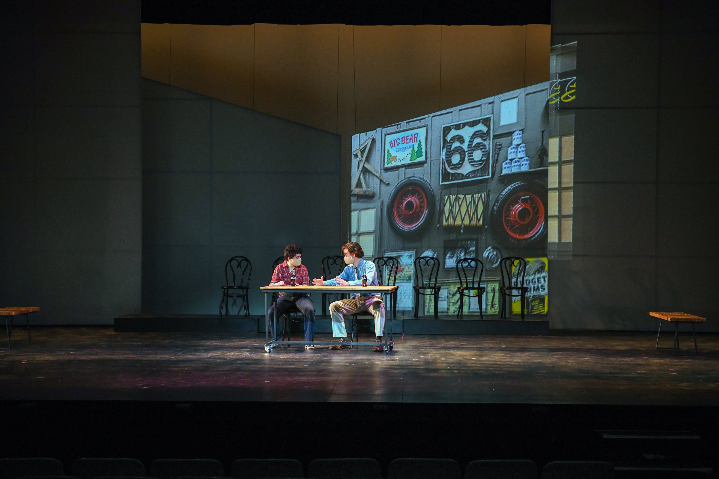Two actors sitting at a table on a theater stage, engaged in conversation, with a large projected image of a vintage store front behind them. The store front has a sign that reads 'Big Bear' and displays various signs, including US Route 66.
