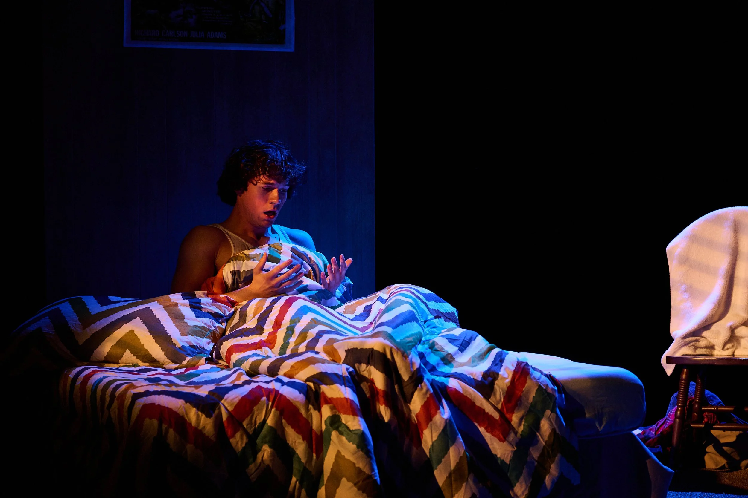 A young person with curly hair is sitting on a bed, looking at their chest with a surprised or shocked expression, in a dimly lit room with a blue hue. The bed has a colorful, zigzag-patterned blanket. There is a framed poster on the wall and a chair