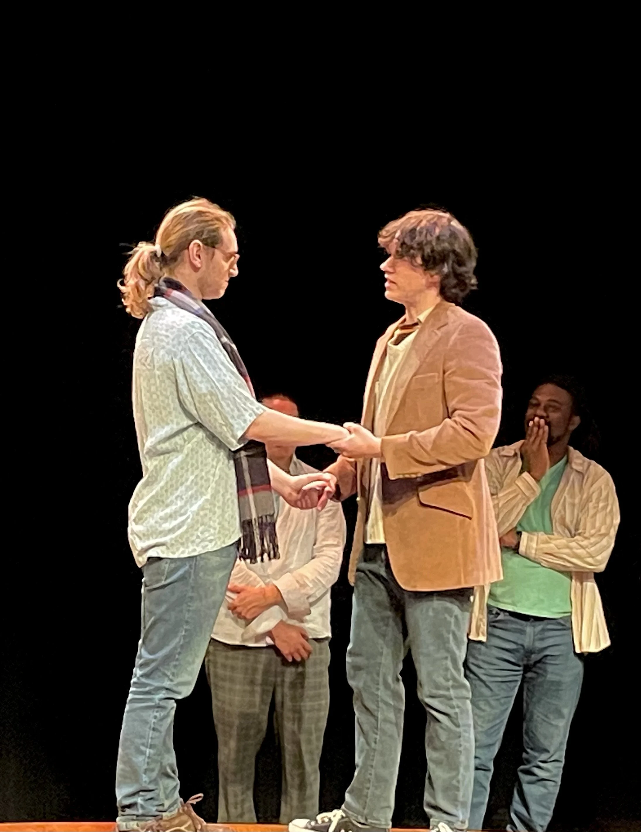 Two men are standing face to face exchanging a handshake or a similar gesture, while other people stand around them on a stage with a black background. The men appear to be engaged in a serious conversation or moment.