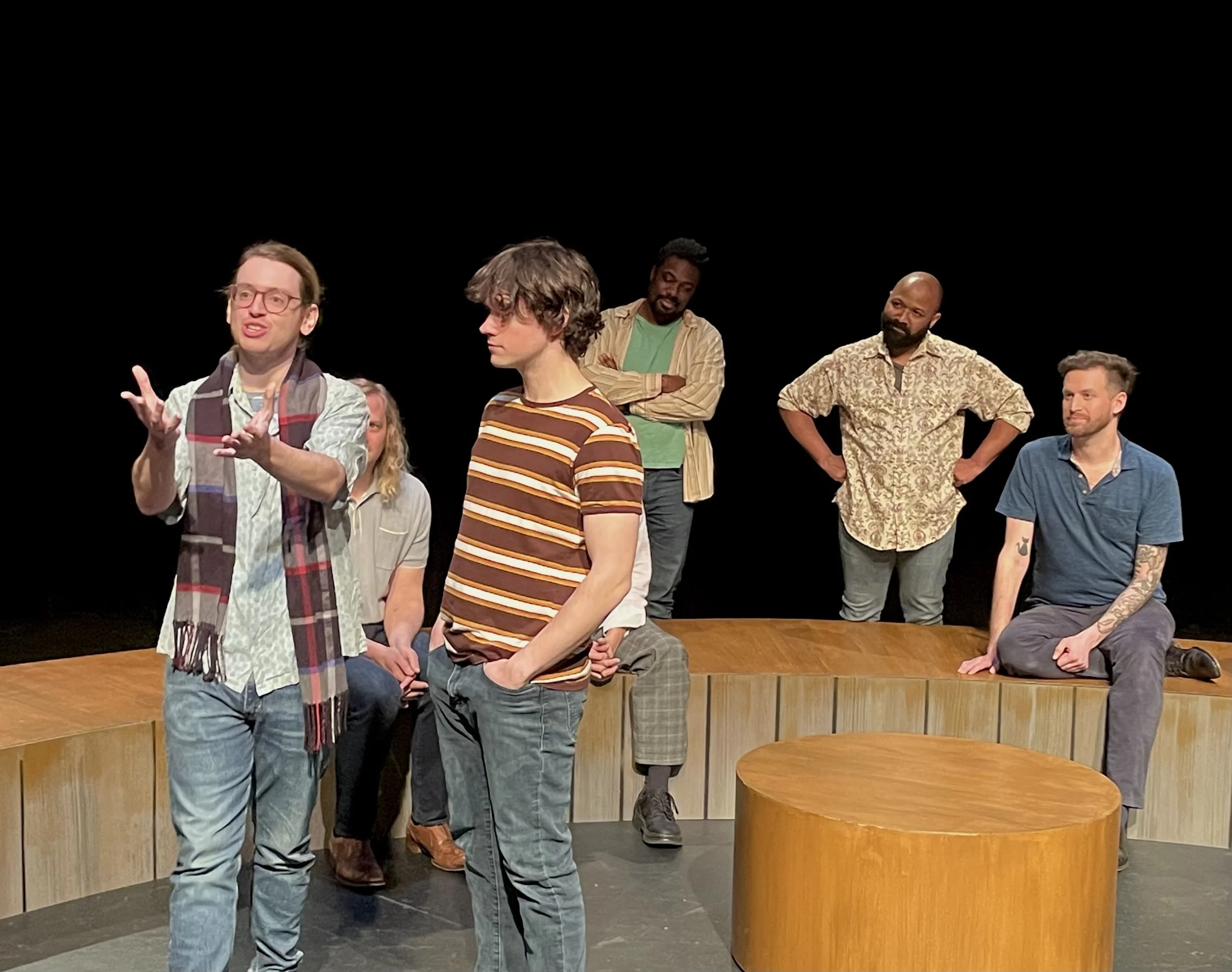A group of people standing and sitting on a stage with a black background, engaging in a discussion or presentation, with some mid-conversation and others listening.