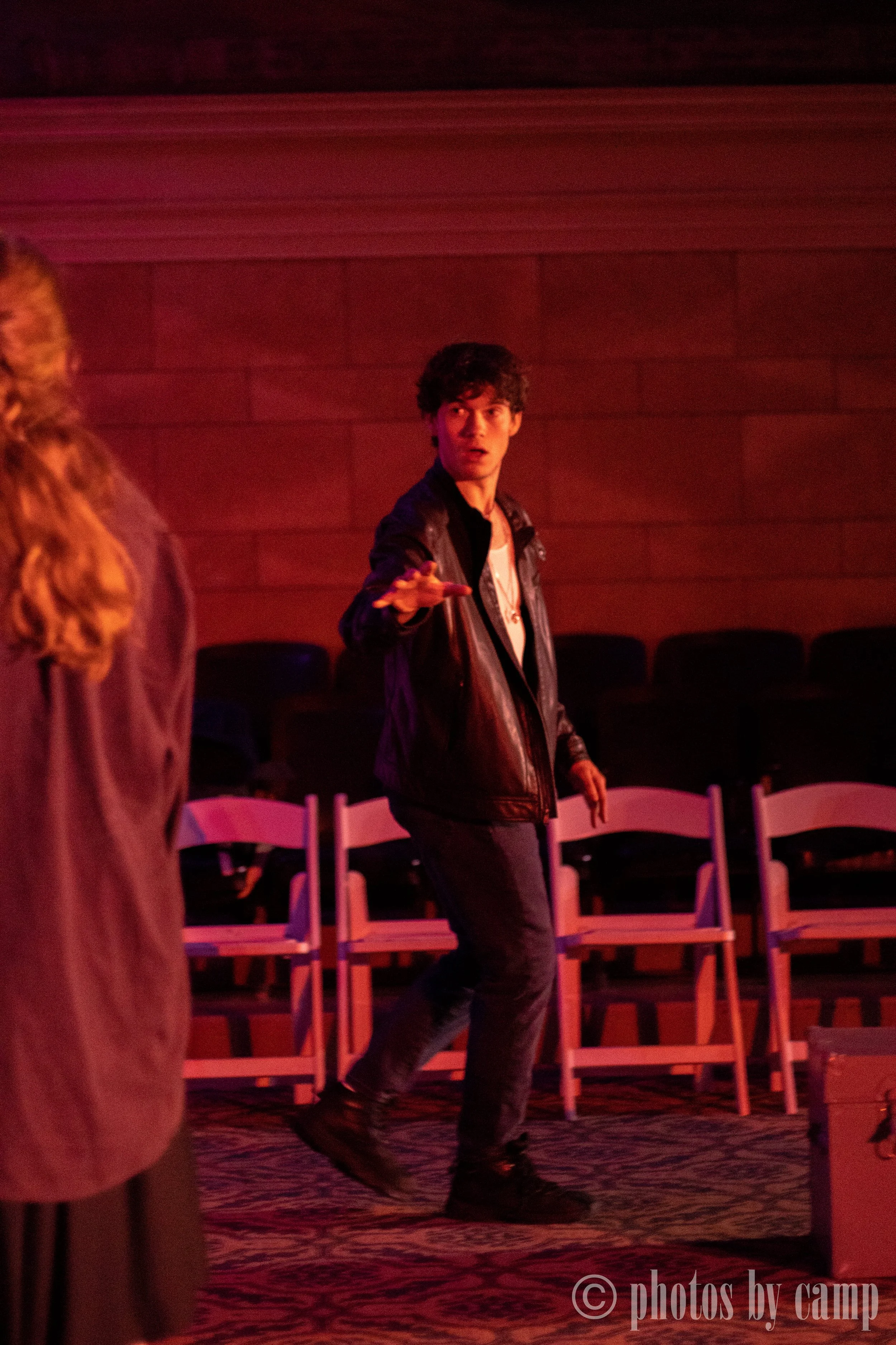 A young man with curly dark hair wearing a black leather jacket and jeans, extending his arm forward in a gesture, standing in a dimly lit room with red and purple lighting, and chairs in the background.
