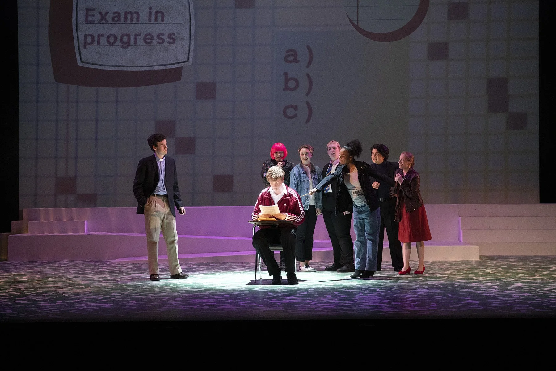 Theater scene with seven actors, some standing and one sitting at a desk, performing on stage with a large projected quiz question in the background.