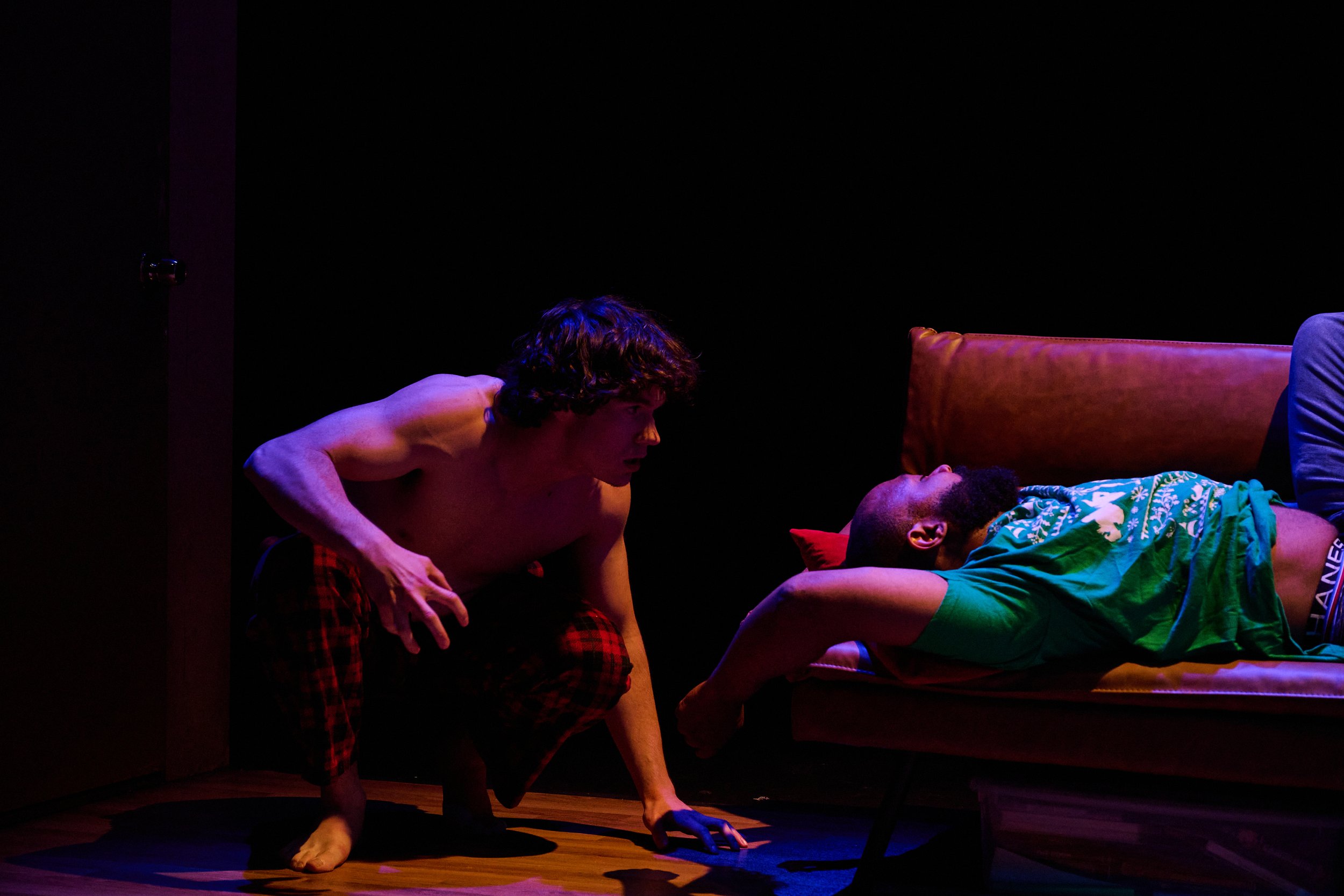 Two men, one shirtless and the other in a green shirt, are on stage with a black background. The shirtless man is leaning toward the man lying on the bed, who is wearing a patterned shirt. There is a red cushion and a partially visible leg on the bed