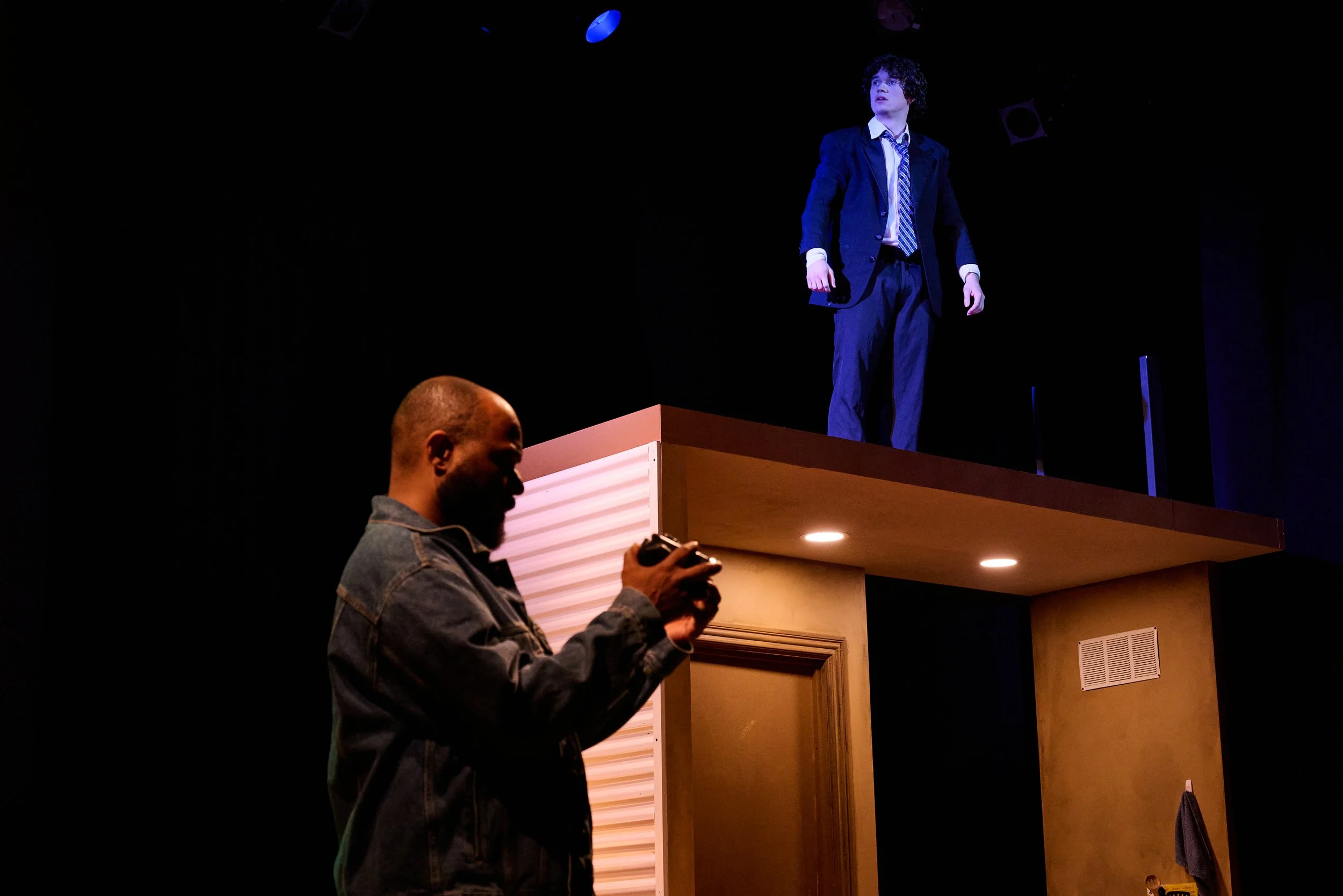 A young man in a dark suit and striped tie stands on a stage, looking upward, with stage lights illuminating him, while a person in a denim jacket is in the foreground, holding a camera or phone, capturing the scene.