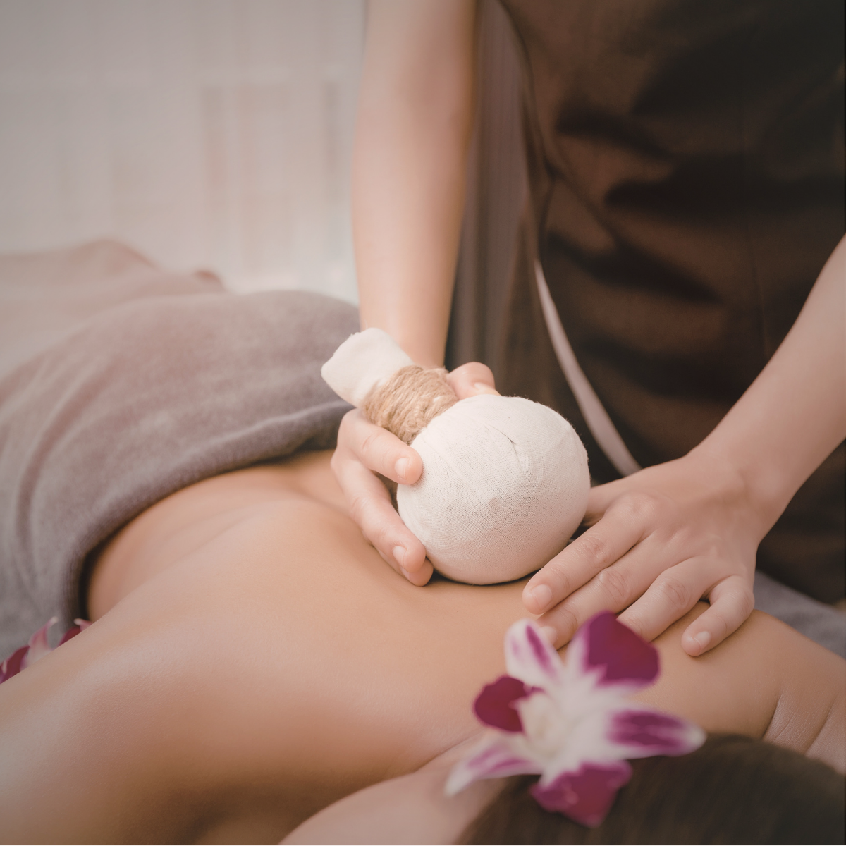A person receiving a massage with a herbal compress on their lower back, while another person’s hand gently supports them; a flower adorns the person's hair.