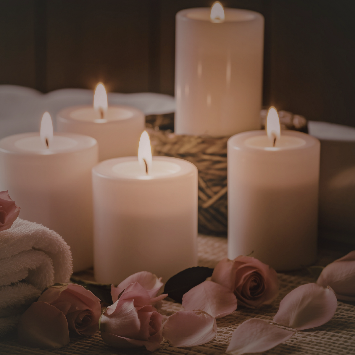 Five lit white candles of varying heights arranged on a table with scattered pink rose petals and a rolled white towel, creating a calming and romantic atmosphere.
