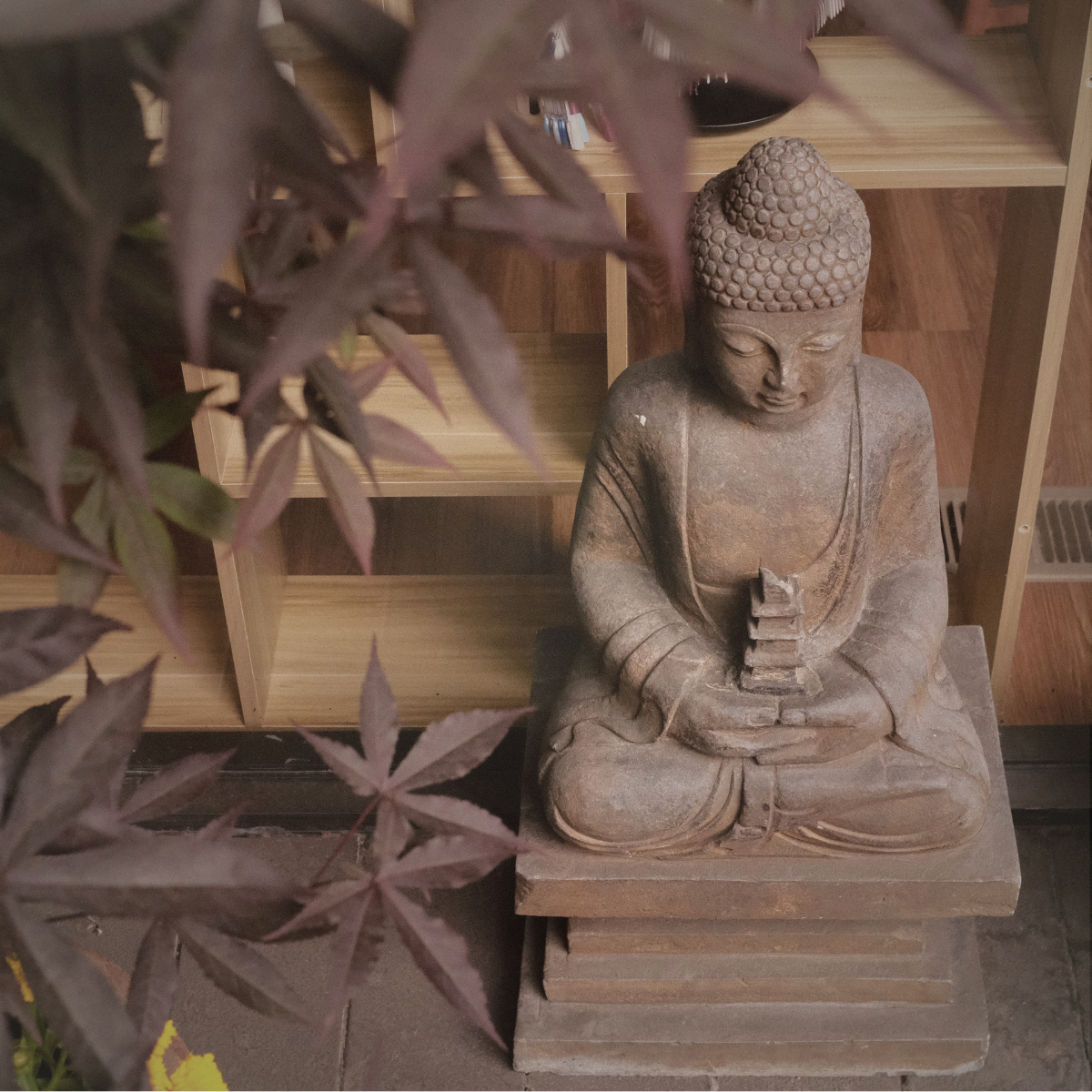 Wooden Buddha statue sitting cross-legged with hands in lap, surrounded by wooden shelves and purple leaves in the foreground.