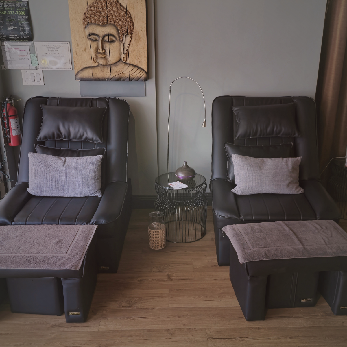 Two black leather massage chairs with grey pillows and towels, positioned side by side in a spa or salon, with a round wireframe side table and modern floor lamp between them, a Buddha artwork on the wall behind, and a fire extinguisher nearby.
