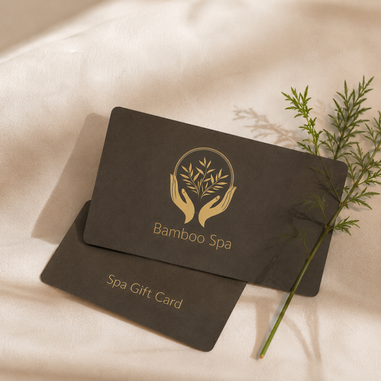 A black spa gift card with gold logo and text for Bamboo Spa, placed on cream-colored fabric, with a sprig of greenery beside it.