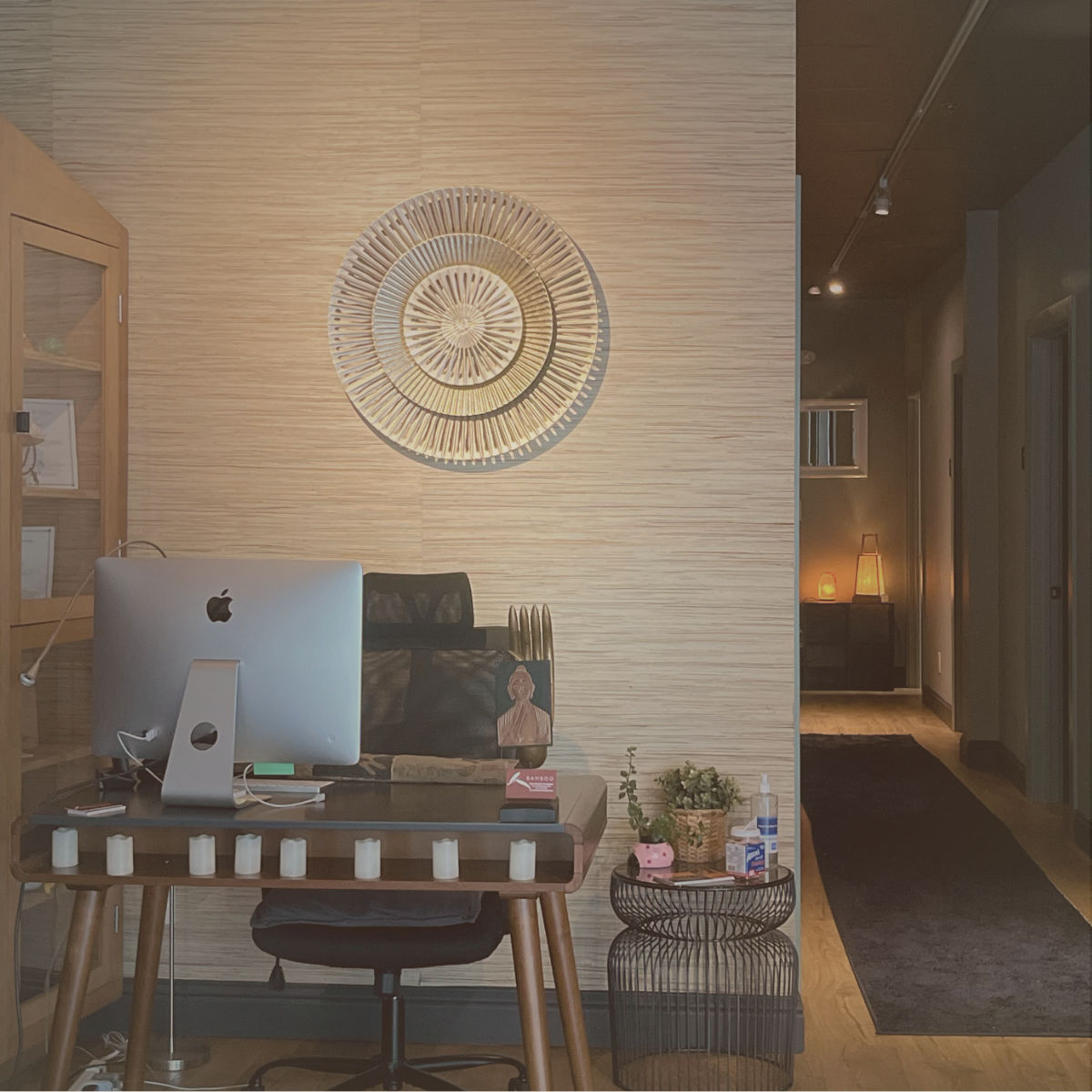 A modern office space with a wooden desk, an Apple computer, black ergonomic chair, decorative candles on the table, and a small side table with plants and hand sanitizer. A decorated wall with a large circular artwork is behind the desk, and a doorway leads to a softly lit hallway with lamps.