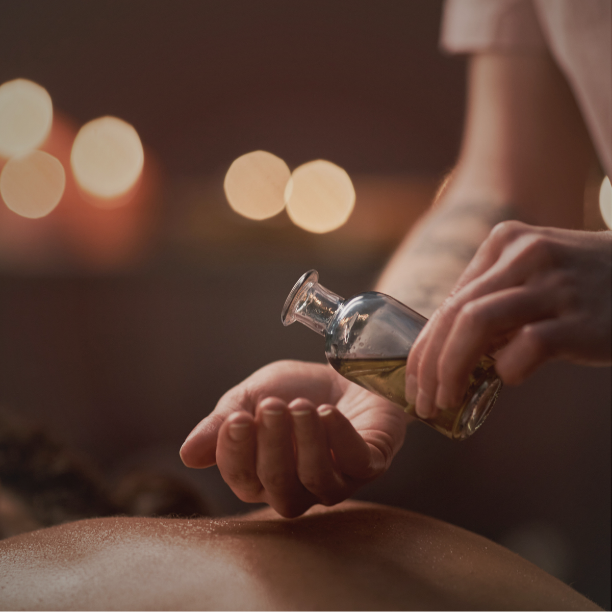 Close-up of a person pouring oil onto another person's back, with blurred warm lights in the background.