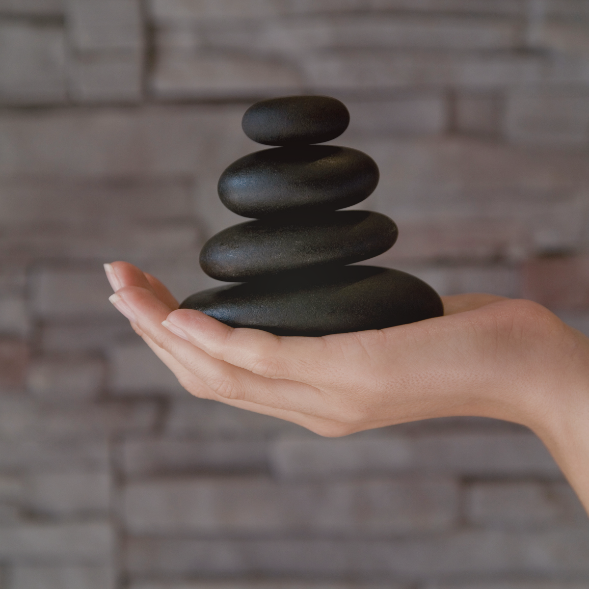 A hand holding a stack of five smooth black stones against a gray stone wall background.