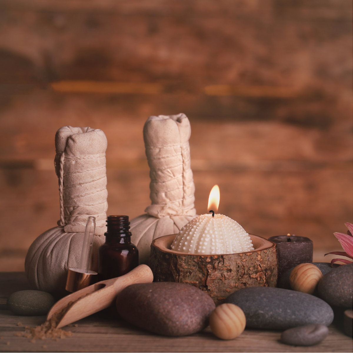 Spa or wellness scene featuring candles, massage herbal packs, stones, and essential oils on a wooden surface.