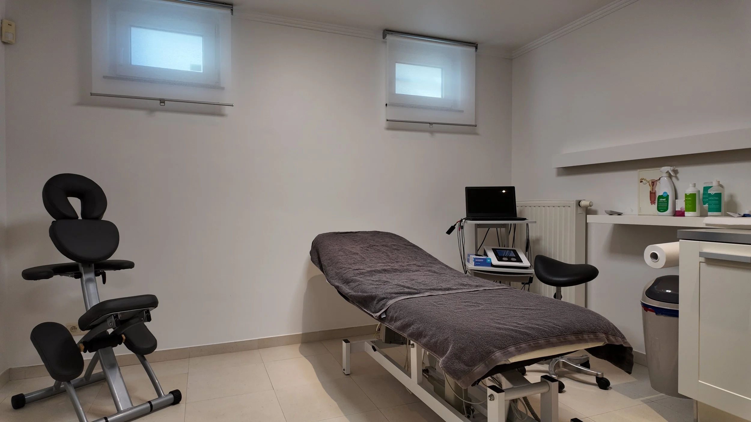 Une salle médicale avec un lit d'examen recouvert d'une couverture grise, une chaise ergonomique noire, un petit bureau avec un ordinateur portable, un appareil médical, et des étagères contenant des bouteilles de produits médicaux et du papier toile