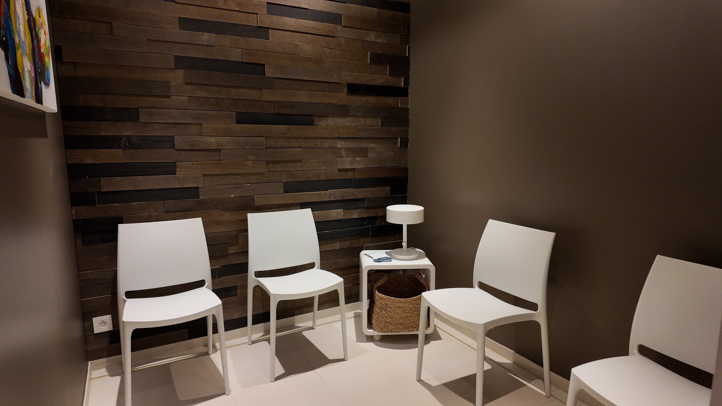 Une salle d'attente avec quatre chaises blanches en plastique, un petit meuble blanc avec une lampe de table et un panier en osier dessous, une paroi en bois foncé et un mur beige