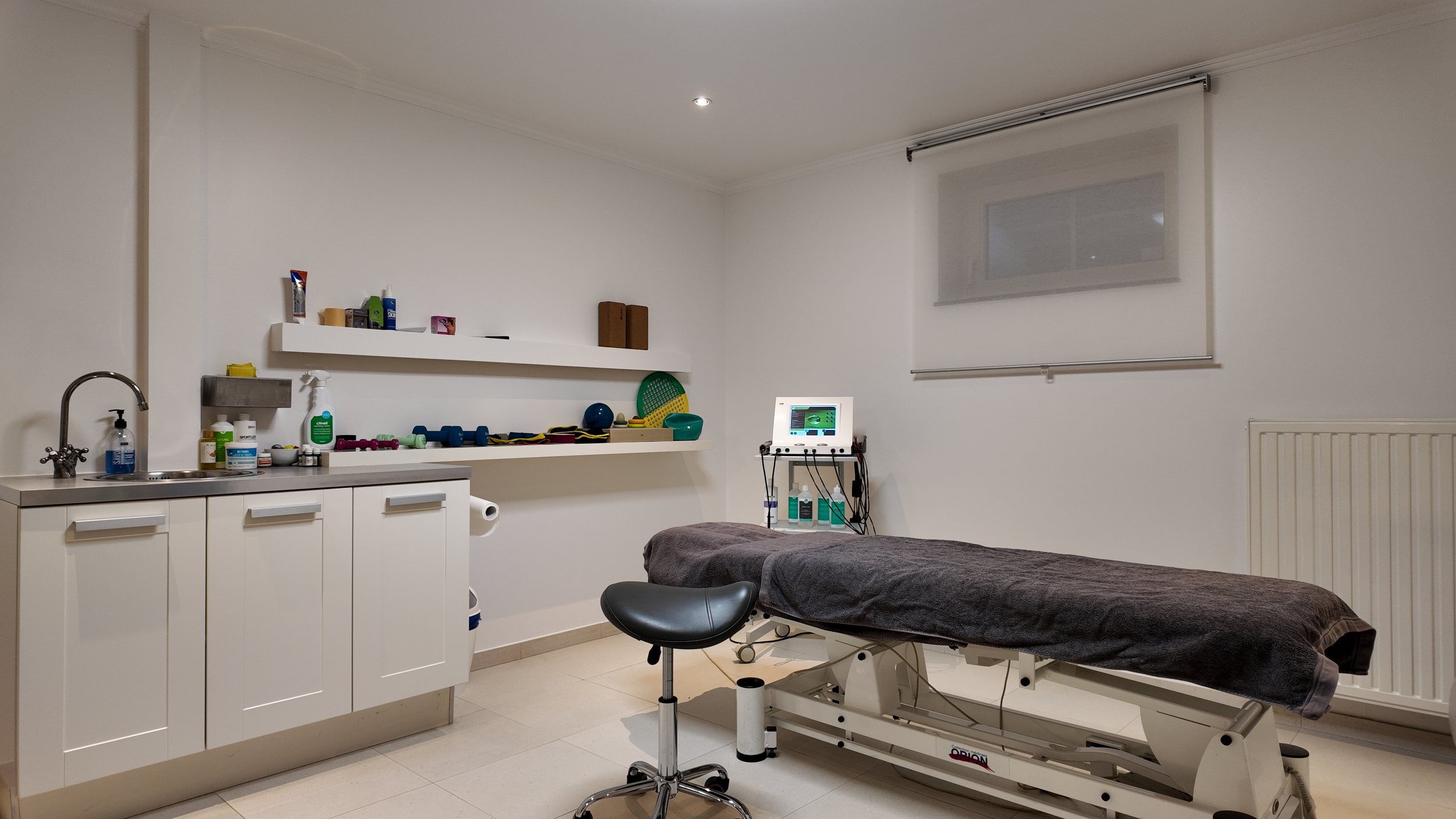 Une salle de consultation médicale avec un lit d'examen, une station d'équipement médical, et des étagères avec des fournitures médicales, dans un environnement clinique propre et bien éclairé.