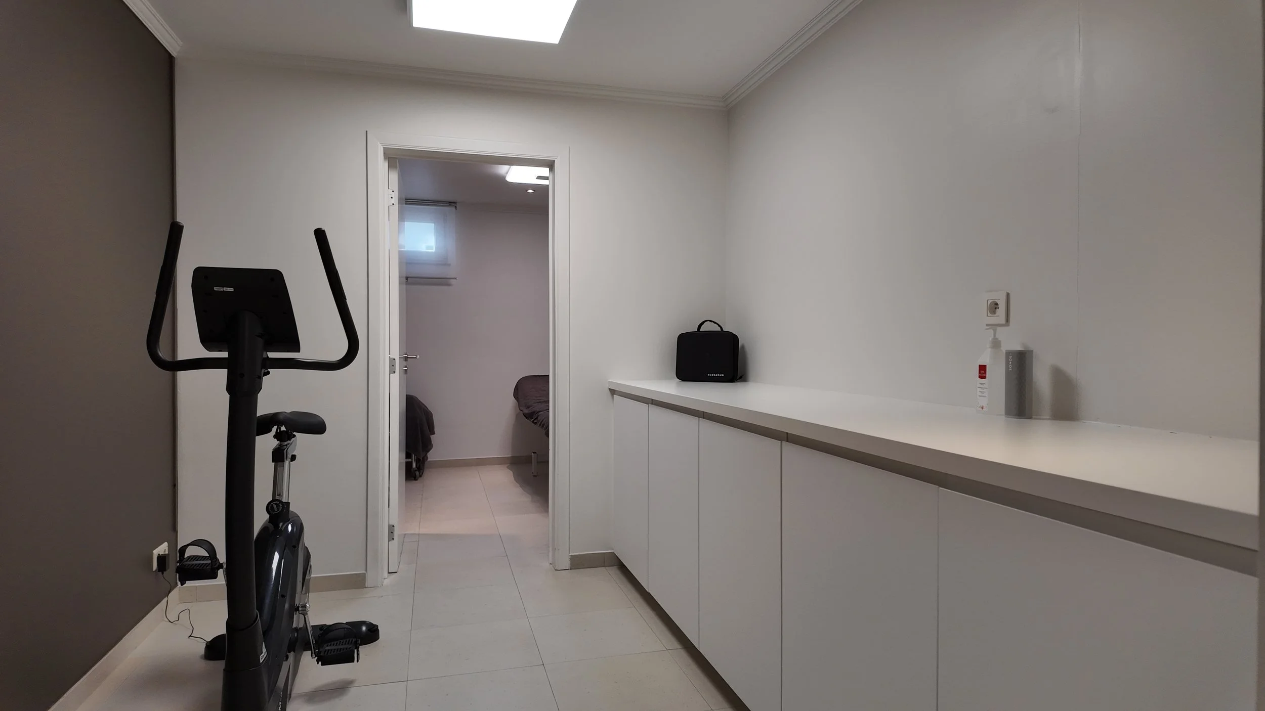 Salle blanche avec un vélo d'appartement, un comptoir blanc avec une enceinte noire, une bouteille de gel désinfectant et une prise électrique, mur blanc, plafond avec éclairage