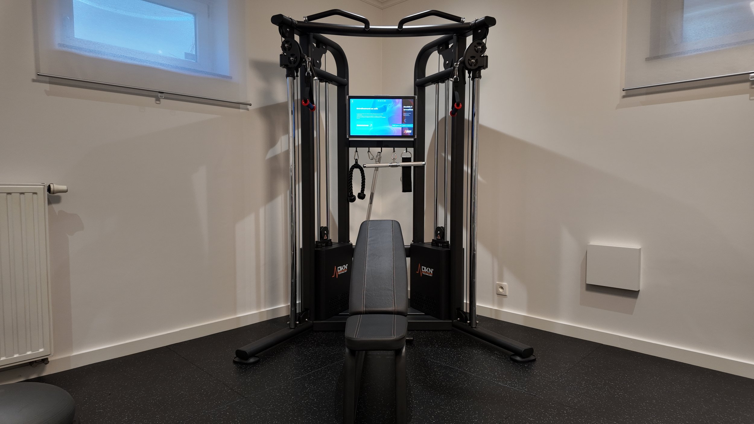 Une machine de musculation dans une salle d'entraînement, équipée de câbles et de pinces de traction, avec un écran au centre et un fauteuil devant.