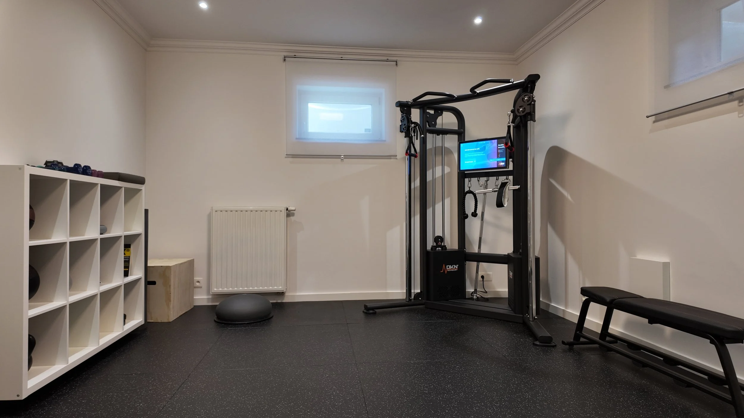 Salle de sport avec équipement de musculation, un petit meuble de rangement, un ballon d'exercice, un banc de musculation, un radiateur blanc, une fenêtre avec voilage blanc, et un mur blanc