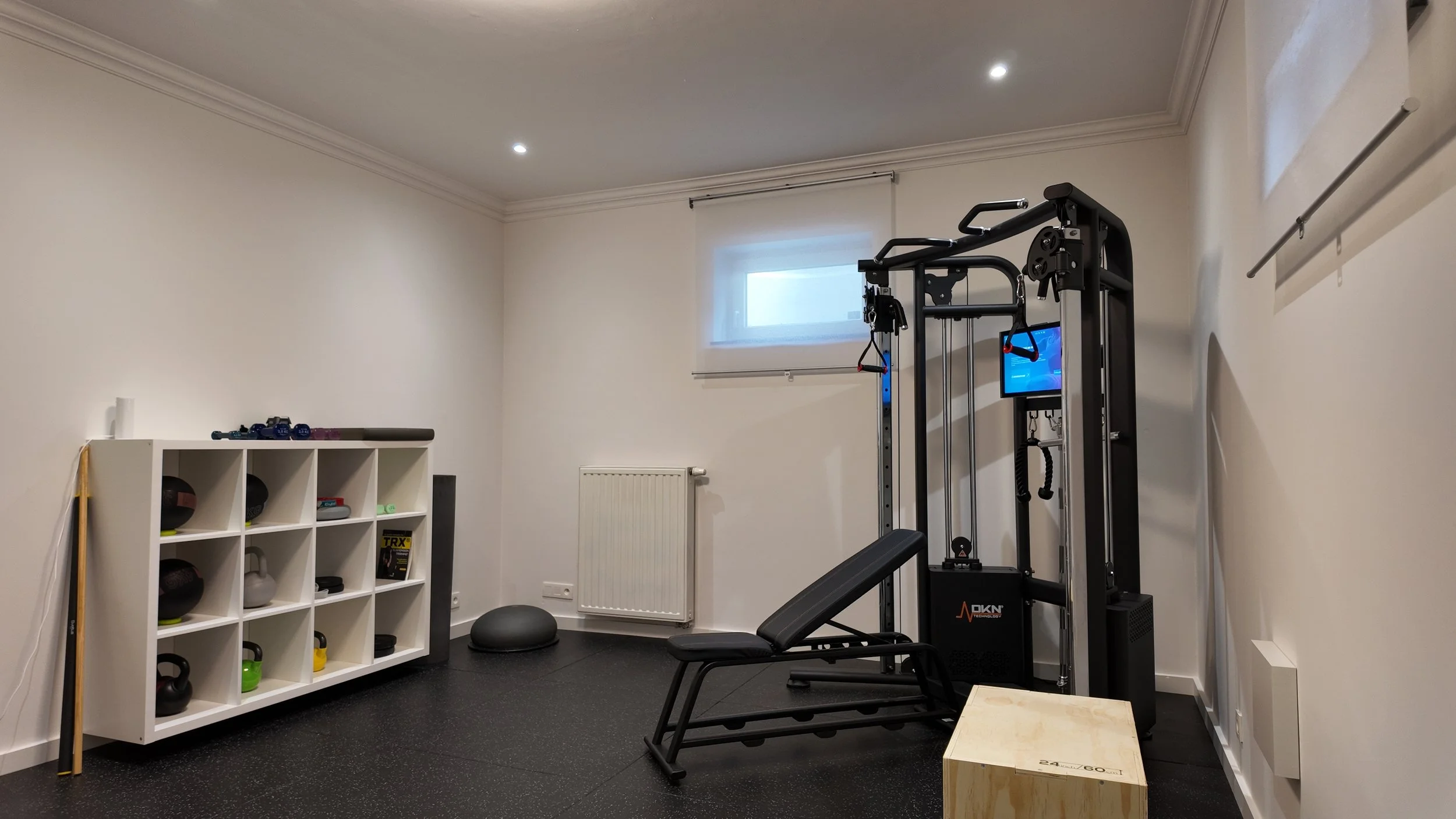 Salle de sport avec un équipement de musculation, un rack d'haltères, et une étagère avec des balles et autres accessoires sportifs.
