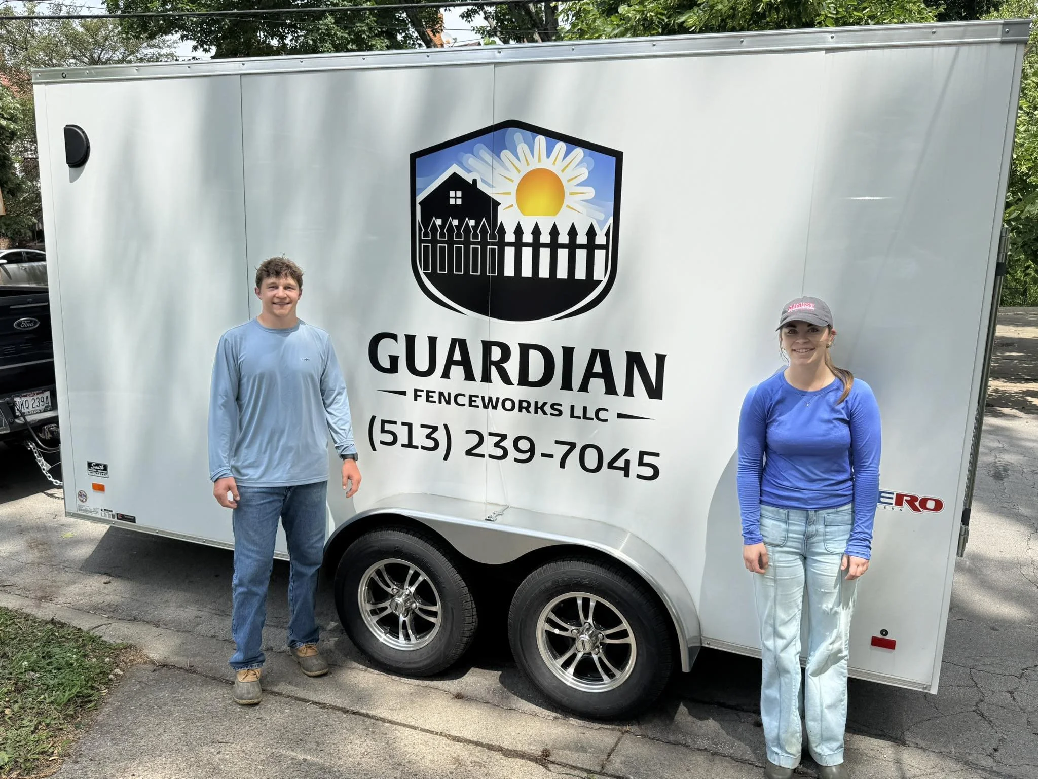 Two people standing in front of a white trailer with a logo and contact information, outdoors under trees. The person on the left is a man wearing a light blue shirt and jeans; the person on the right is a woman in a blue long sleeve shirt, jeans, and a gray cap.