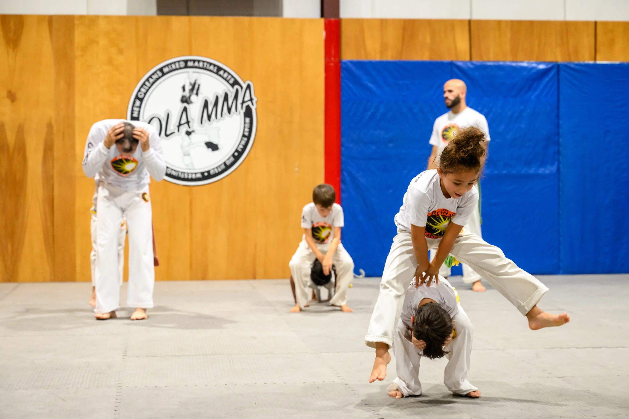 Children practicing martial arts in a dojo, with some standing and others in a piggyback position, wearing white uniforms with a logo featuring a sun and black writing, in front of a wooden wall with a black and white logo for a martial arts academy.