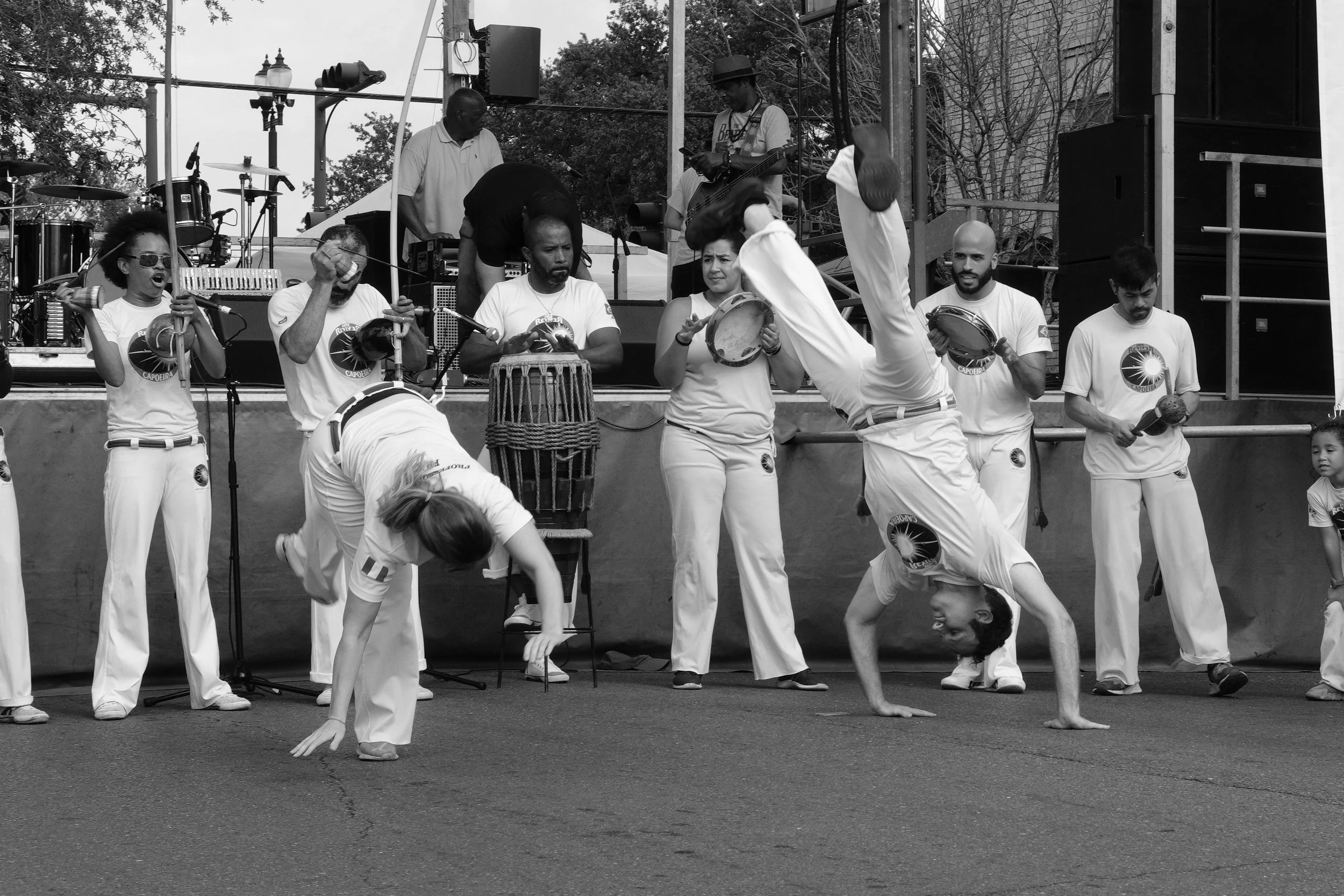 A group of performers on stage involved in a Capoeira dance and martial arts demonstration, some playing instruments, with a musician holding a guitar and others using drums, in an outdoor setting.