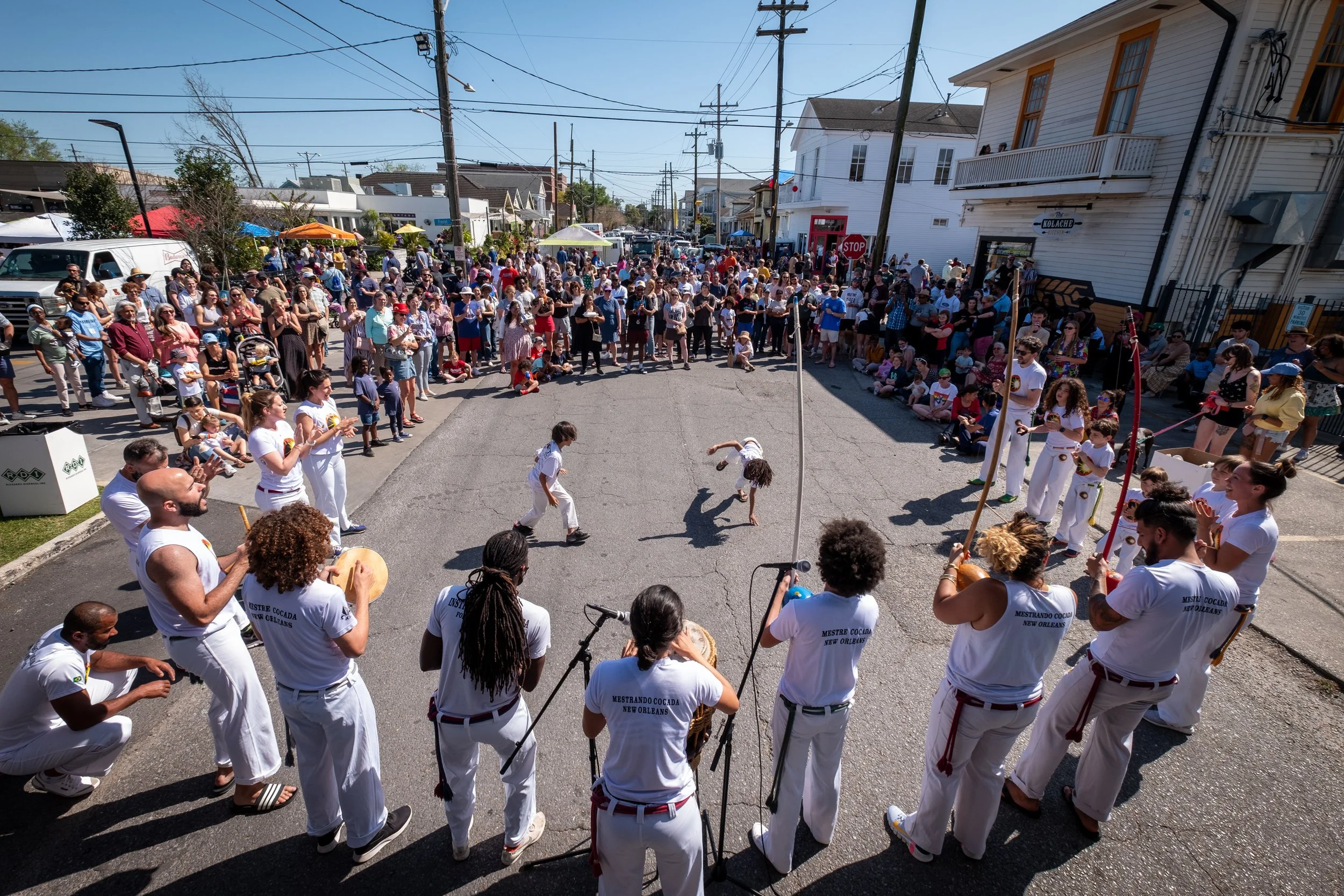 A crowd gathered on a city street during a festival or parade, with people sitting and standing on sidewalks, some performing capoeira, and many holding drinks or taking photos. There are colorful umbrellas, a white building with a balcony, and utili