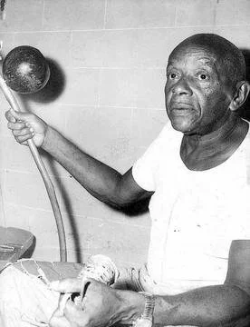 Mestre Bimba holding a berimbau upside-down while seated, wearing a white shirt.