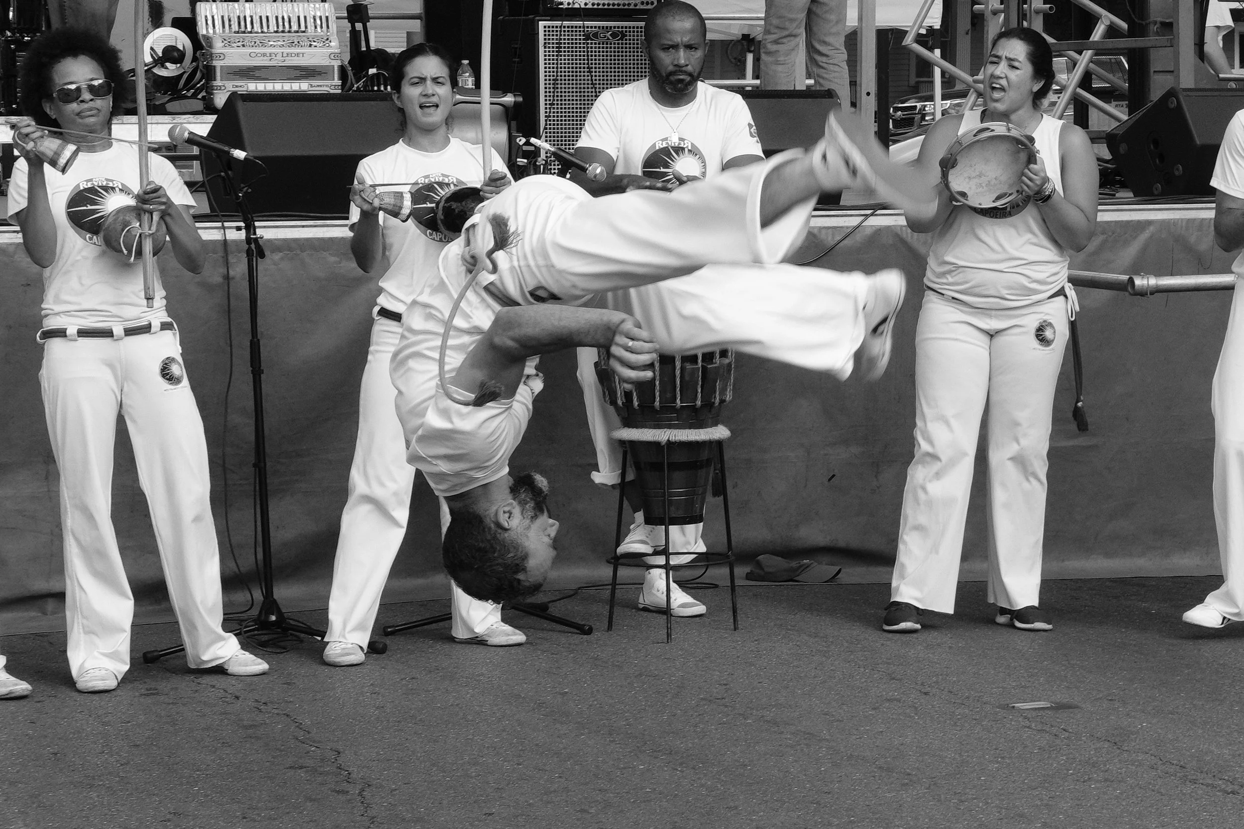 A group of four people, three women and one man, standing on a stage, with one woman performing a capoeira move, upside down. The women are holding percussion instruments, and there are microphones and audio equipment around them.
