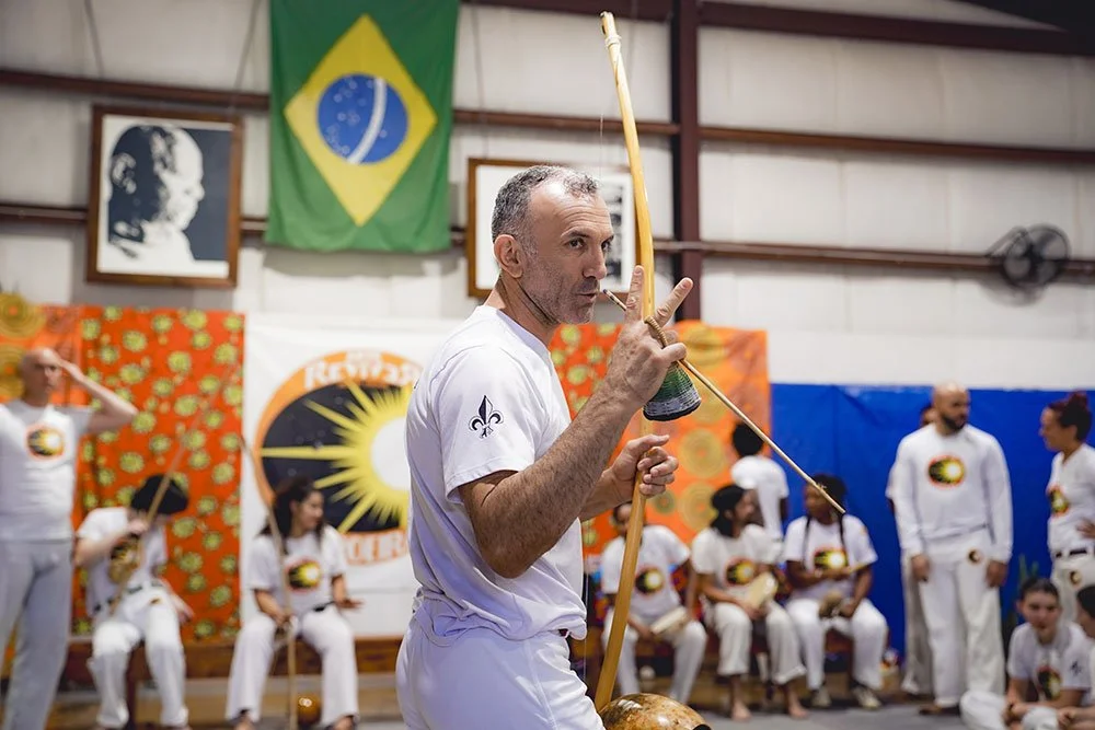 A man in a white shirt playing a berimbau, a Brazilian musical instrument, during a capoeira event. Several people in white clothing sit and stand in the background. The Brazilian flag and other colorful banners decorate the background.