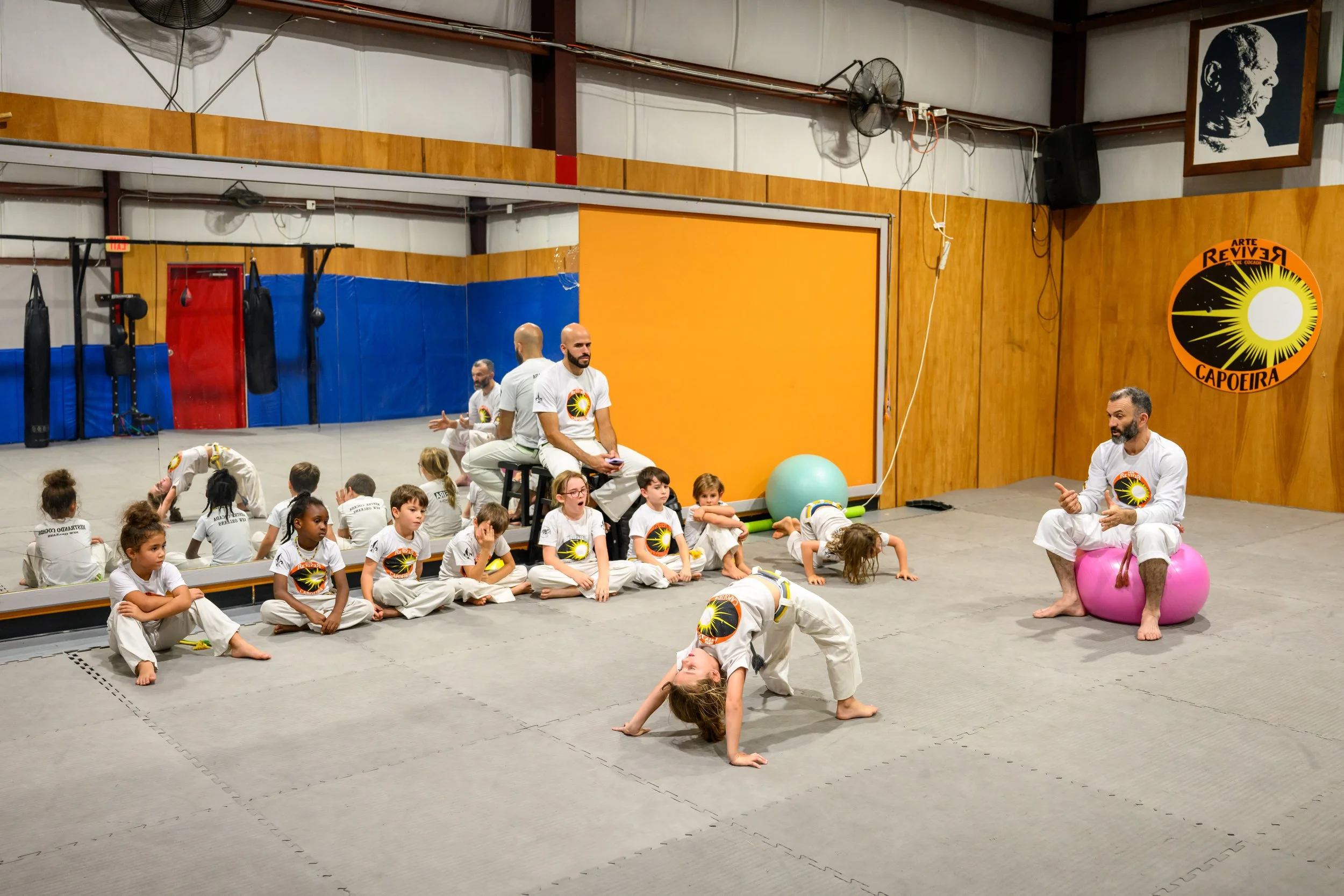 Group of children and instructors practicing capoeira in a gym with wooden walls, punching bags, and a mirror.